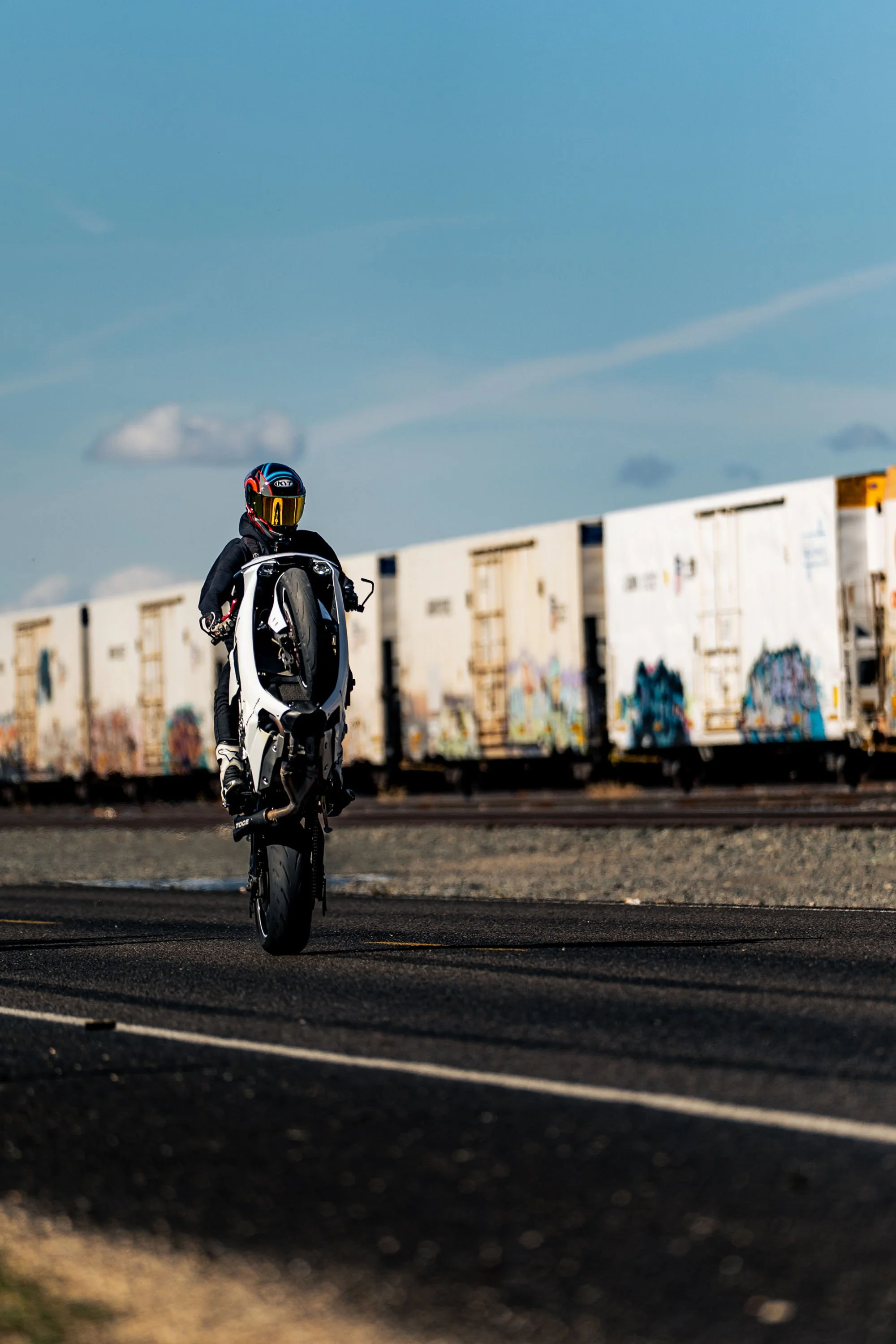 A person riding a white motorcycle on a dark road at sunset near abandoned train cars covered in graffiti.