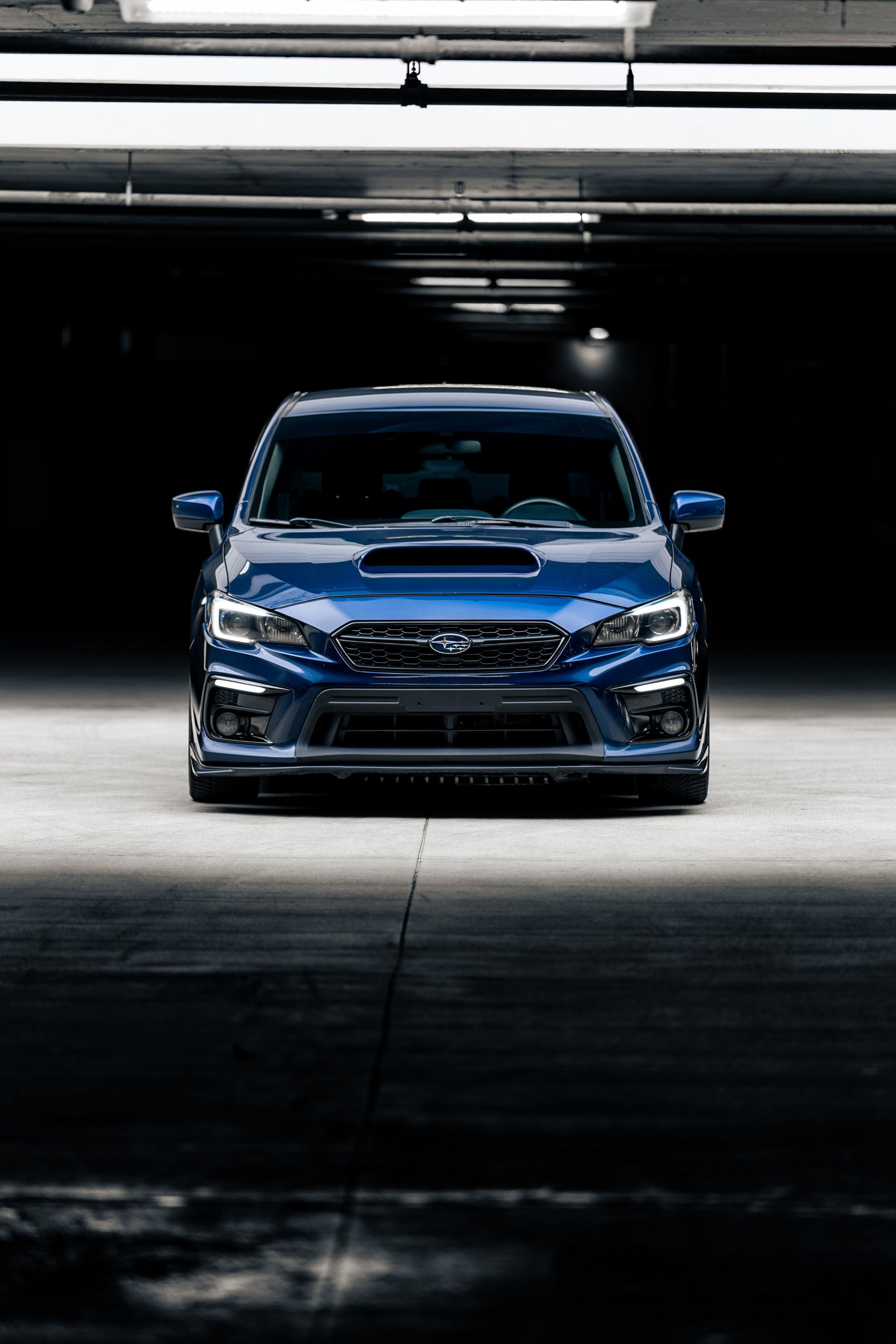 Front view of a blue Subaru car parked in a dark parking garage with overhead lights casting reflections on the car's hood and windshield.