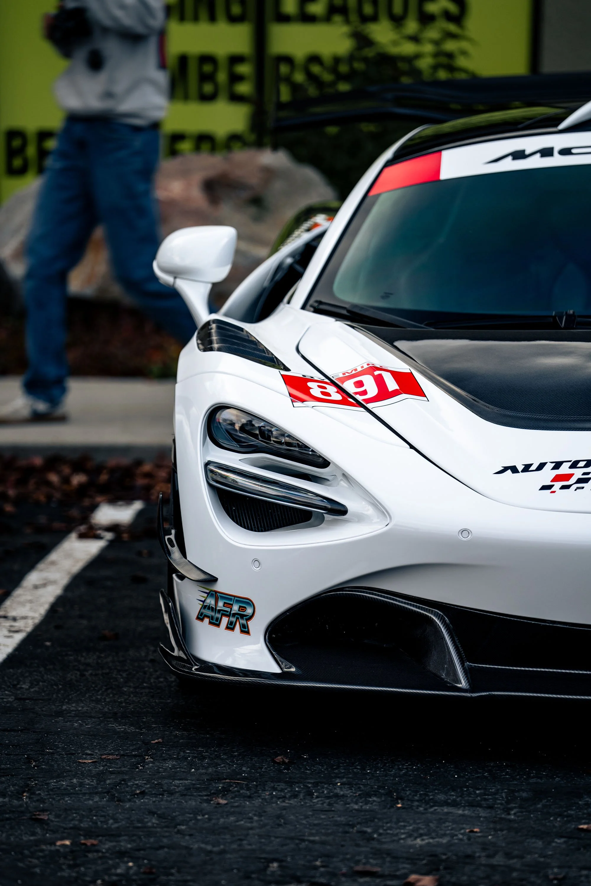Close-up of the front of a white race car with black and red decals, including the number 16. The background features a yellow sign and a person wearing blue jeans and a gray hoodie, slightly out of focus.