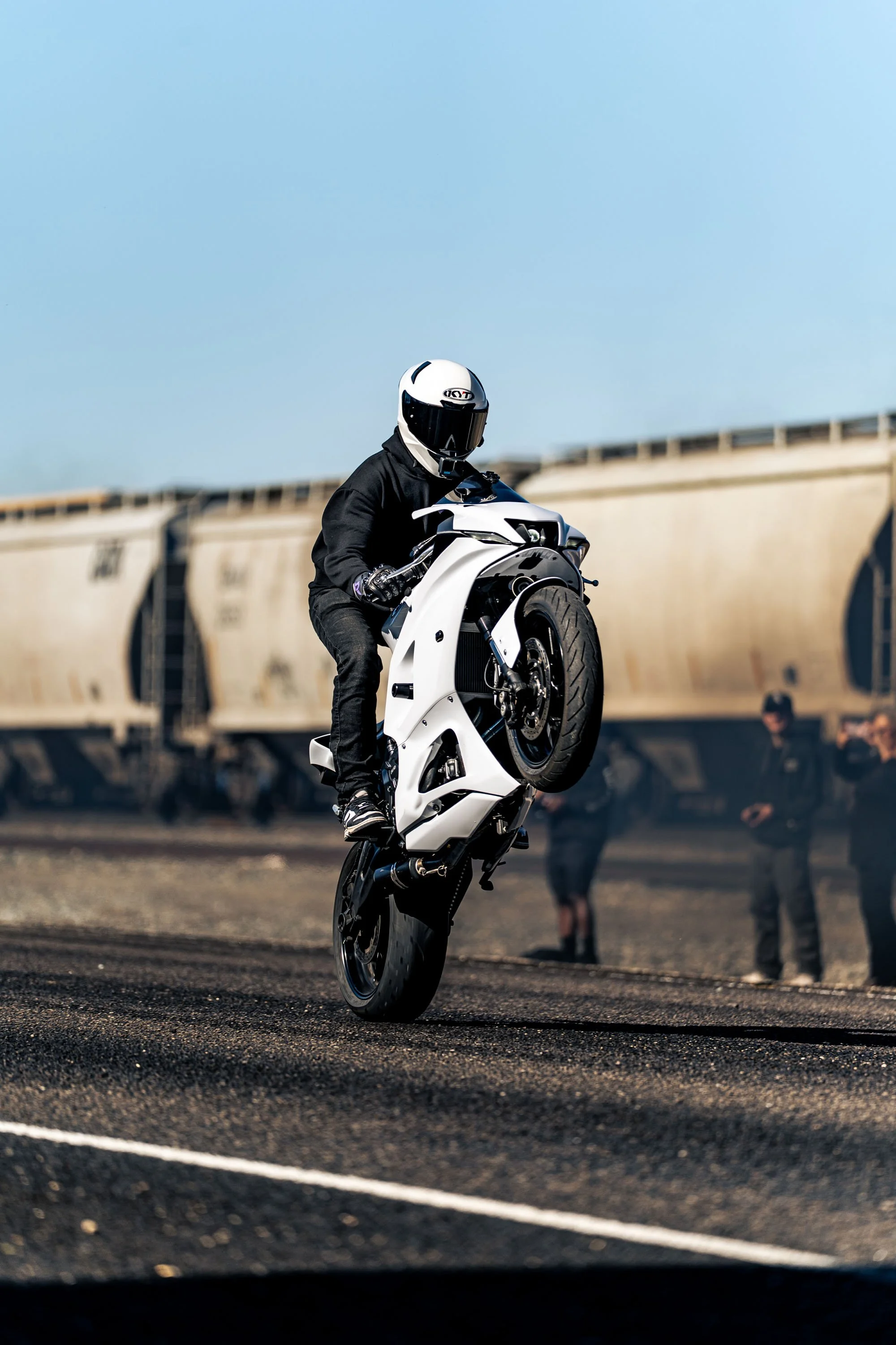 A motorcyclist doing a stunt on a white motorcycle, wearing a black jacket and a white helmet, with people watching in the background at an outdoor location.