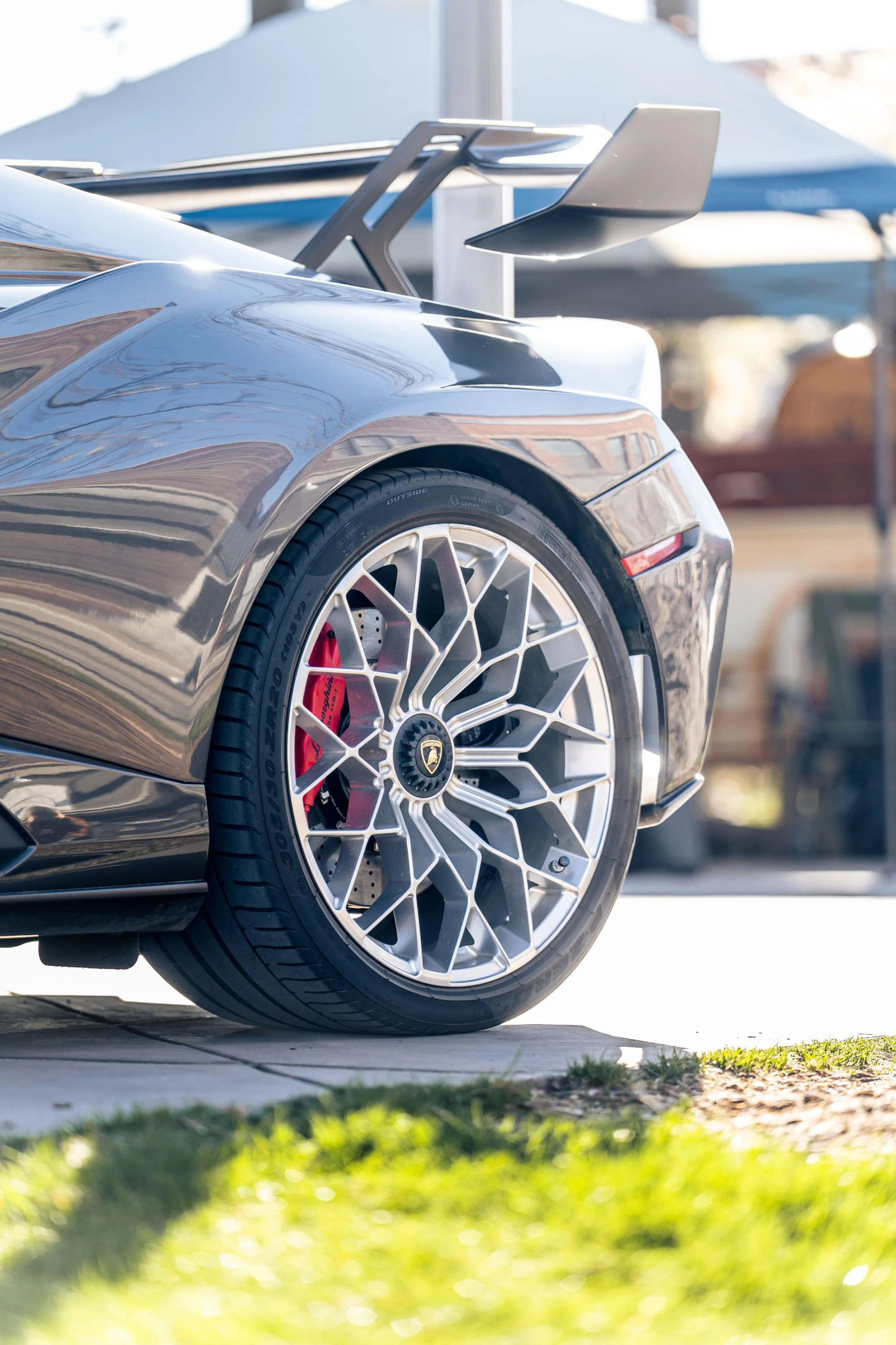 Close-up of the rear wheel of a race car with a distinctive honeycomb rim design, red brake caliper, and slick tire, parked outside near a tent and blurred background.