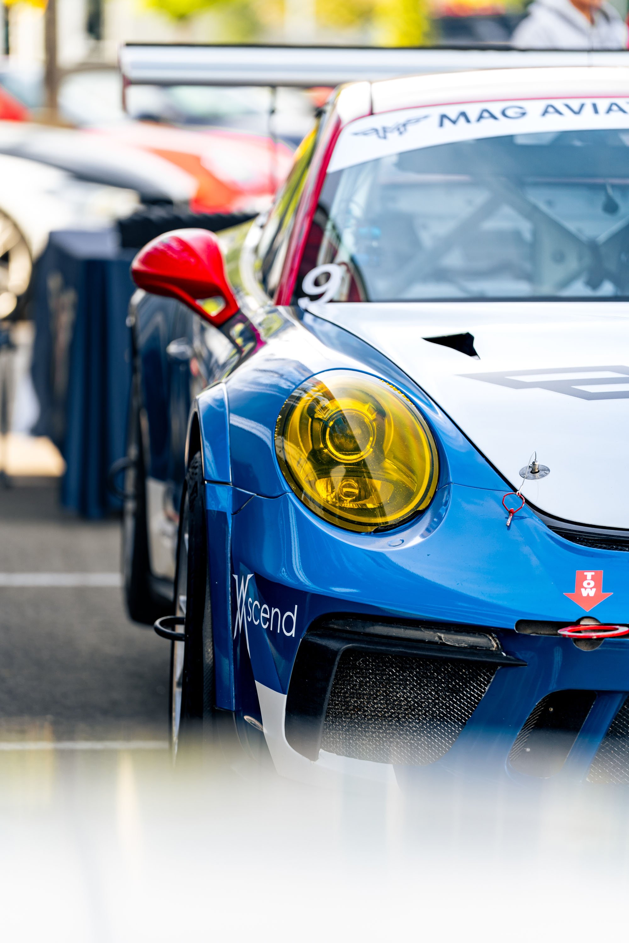Close-up of a blue race car with yellow headlights, red side mirror, and various sponsor logos, parked among other race cars at a racing event.