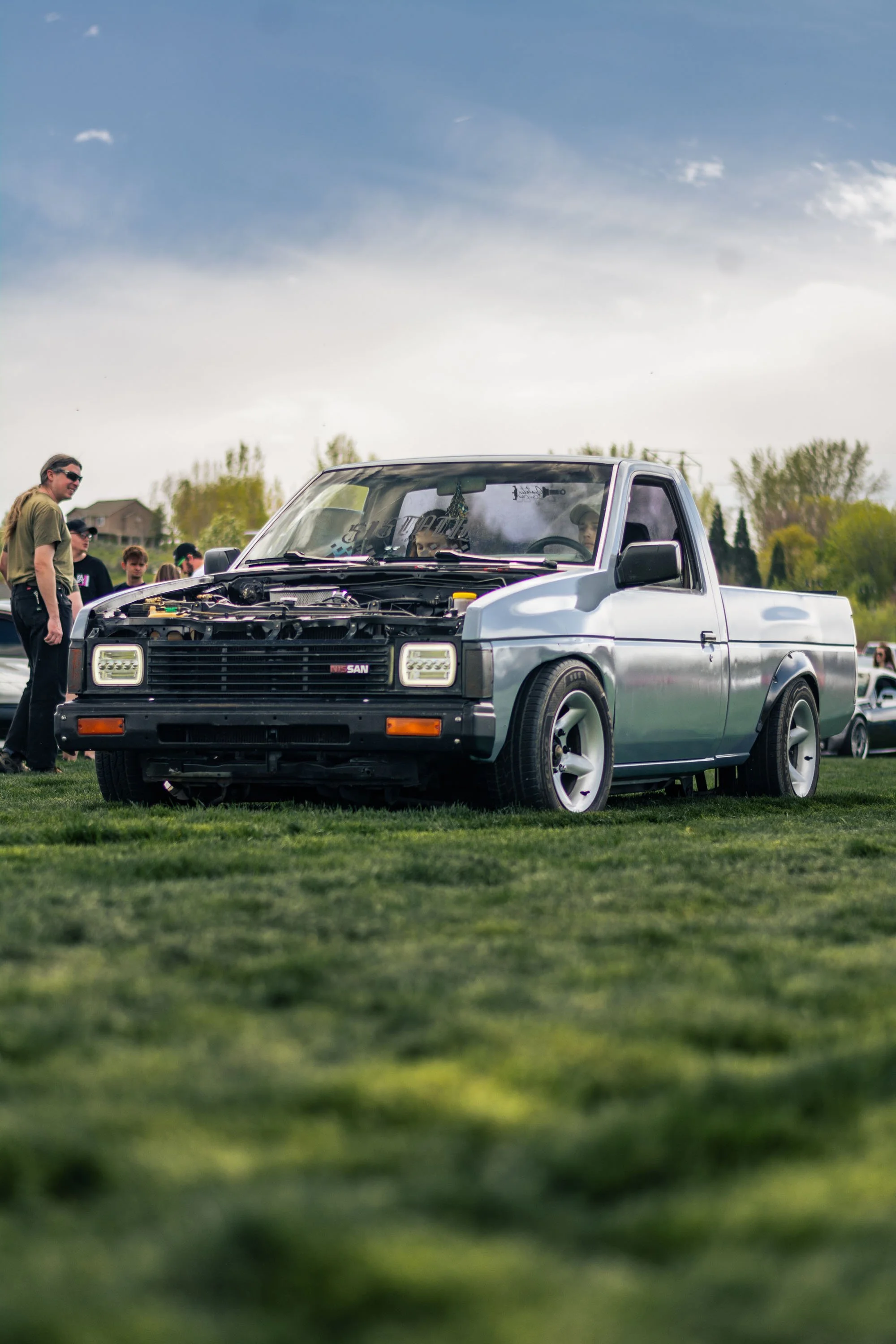 A silver pickup truck with an open hood, parked on grassy field during daytime. Several people are standing nearby, some looking at the engine, others talking. Houses and trees are in the background under a cloudy sky.
