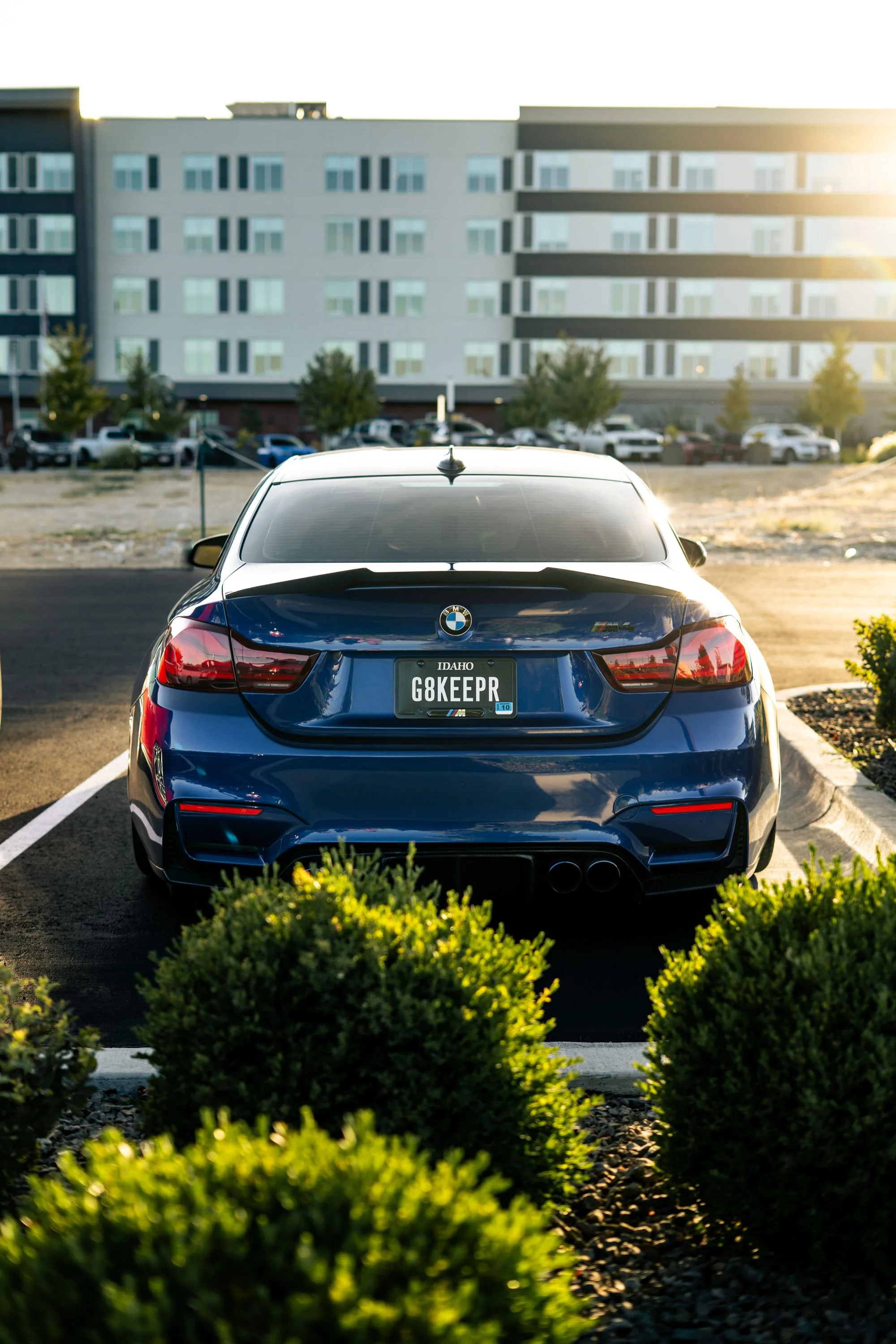 Back of a blue BMW M4 car with Idaho license plate G8KEEPR parked in a parking lot, with a multi-story building and trees in the background, during sunset.