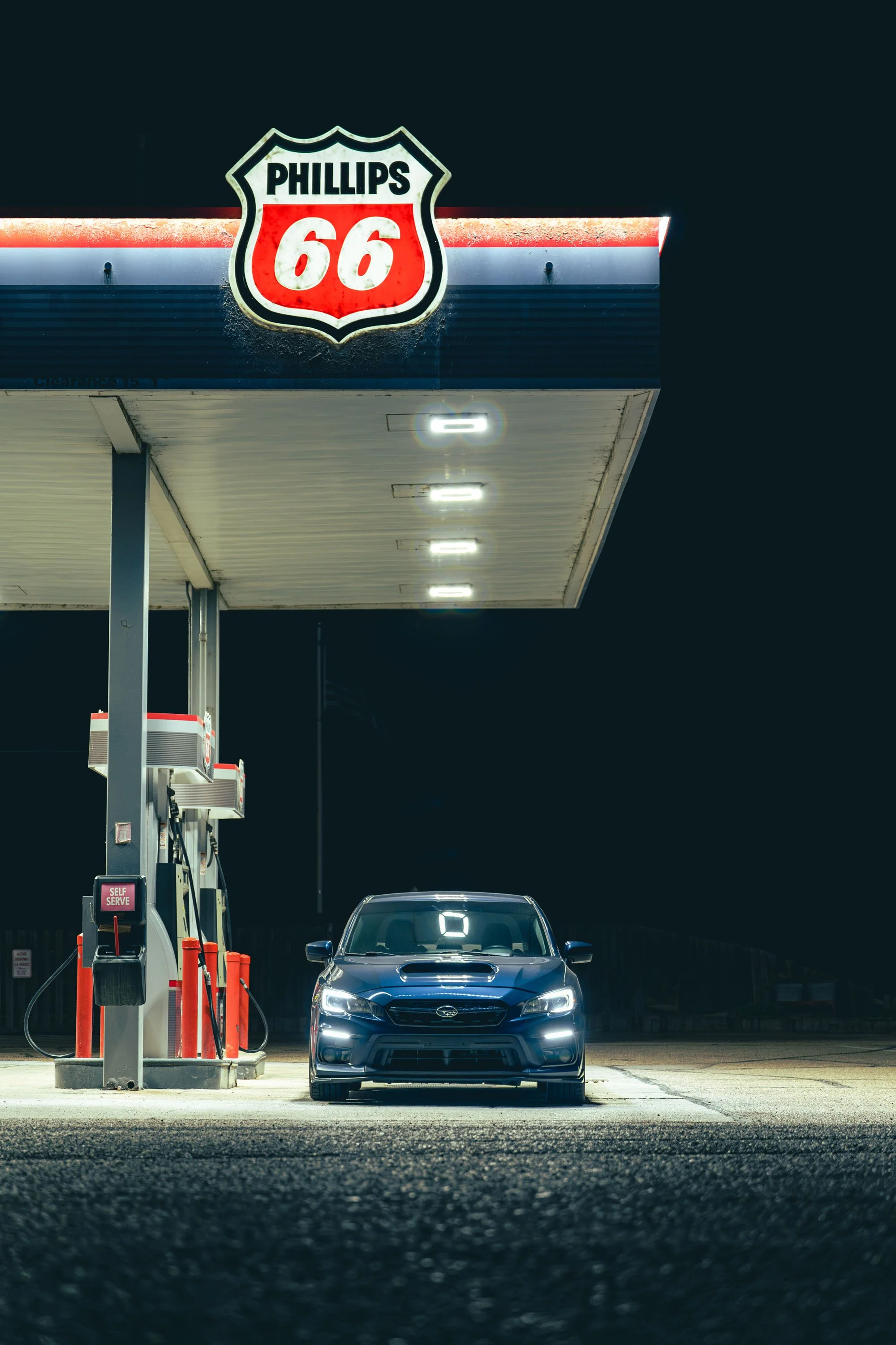 A dark blue sedan parked at a gas station under nighttime lighting, with a Phillips 66 sign visible above.