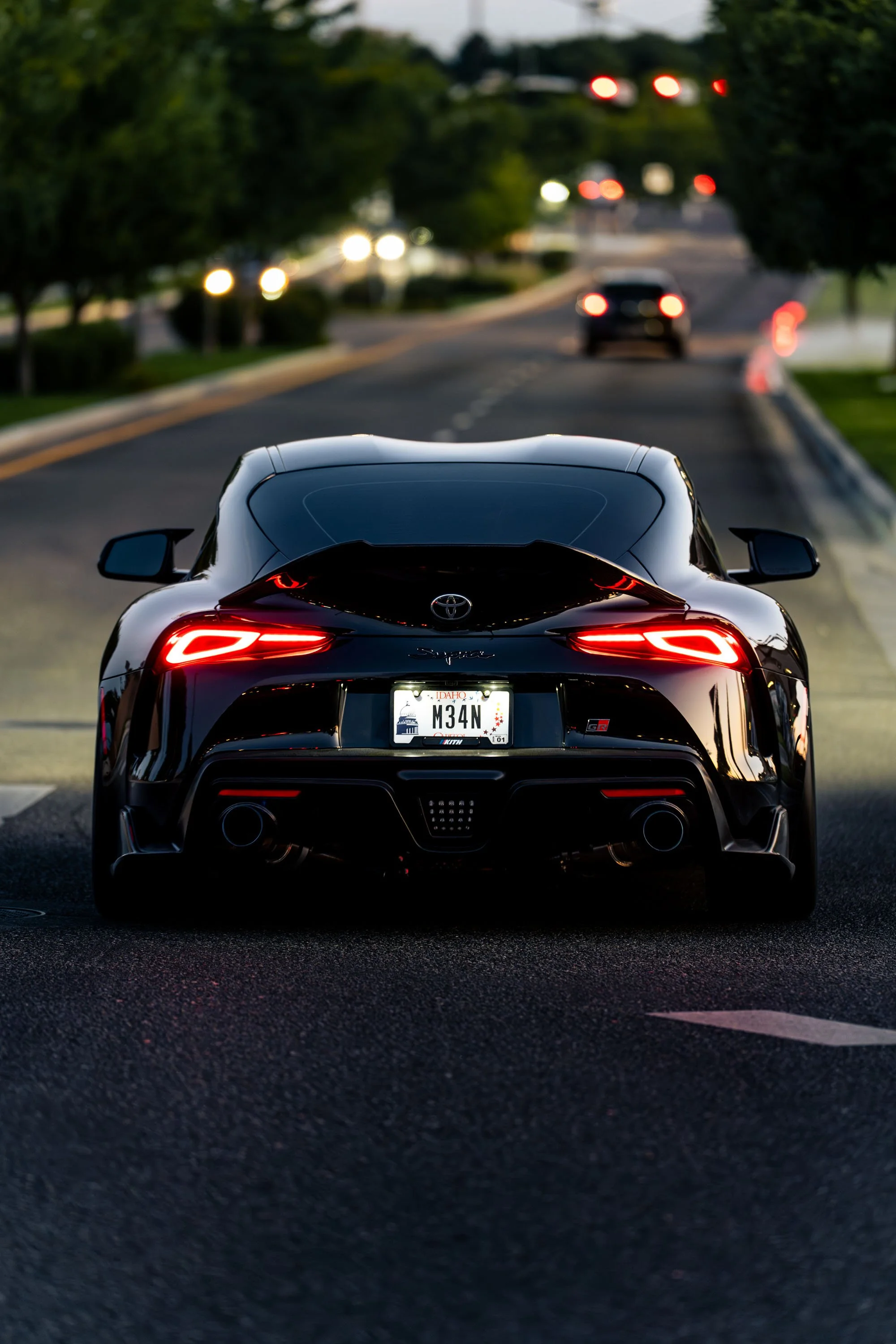 Rear view of a black Toyota sports car with distinctive taillights, parked on a street during dusk or evening, with blurred cars and greenery in the background.