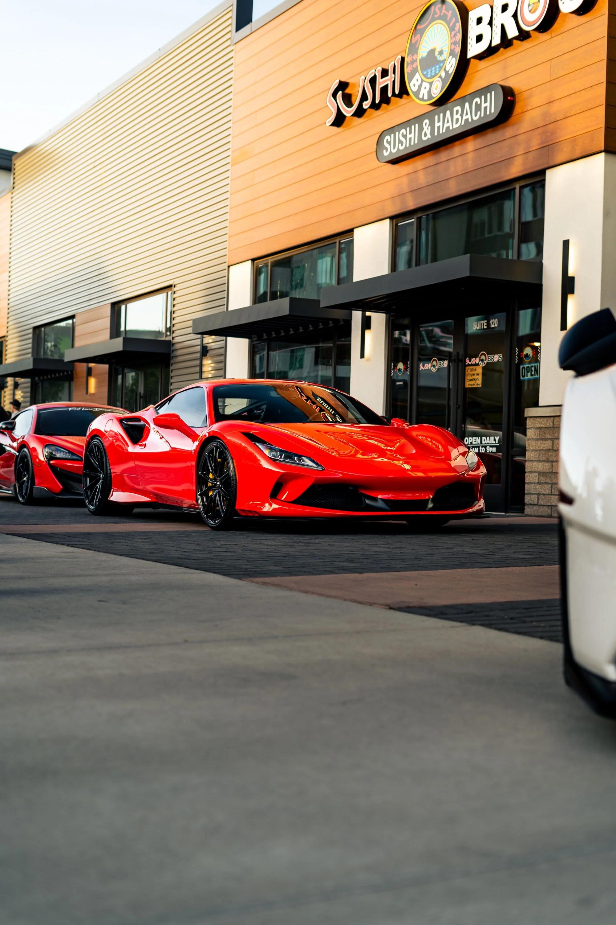 Red sports car parked in front of a sushi restaurant with wooden exterior and sign that reads 'Sushi Bros'.