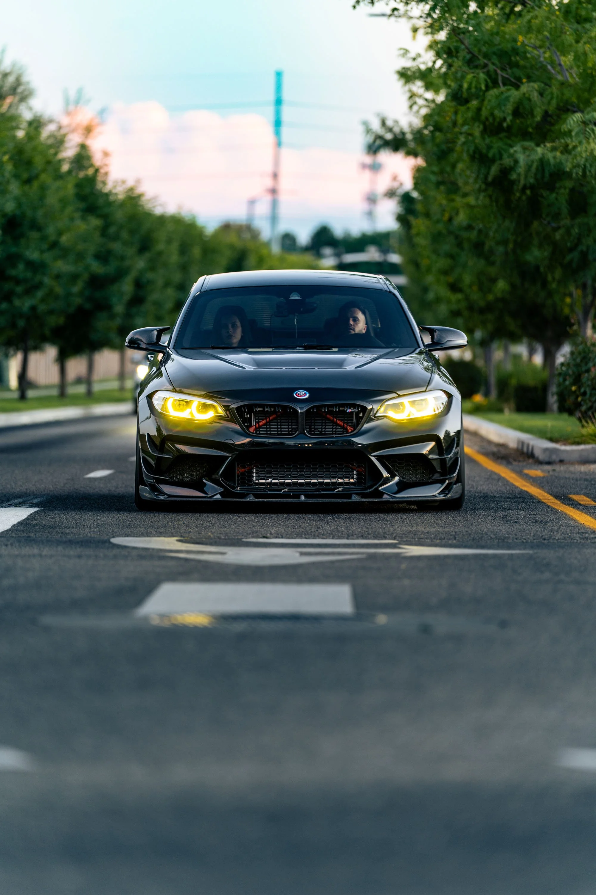 A black sports car with yellow headlights driving on a suburban street lined with green trees, with two people visible inside the car.