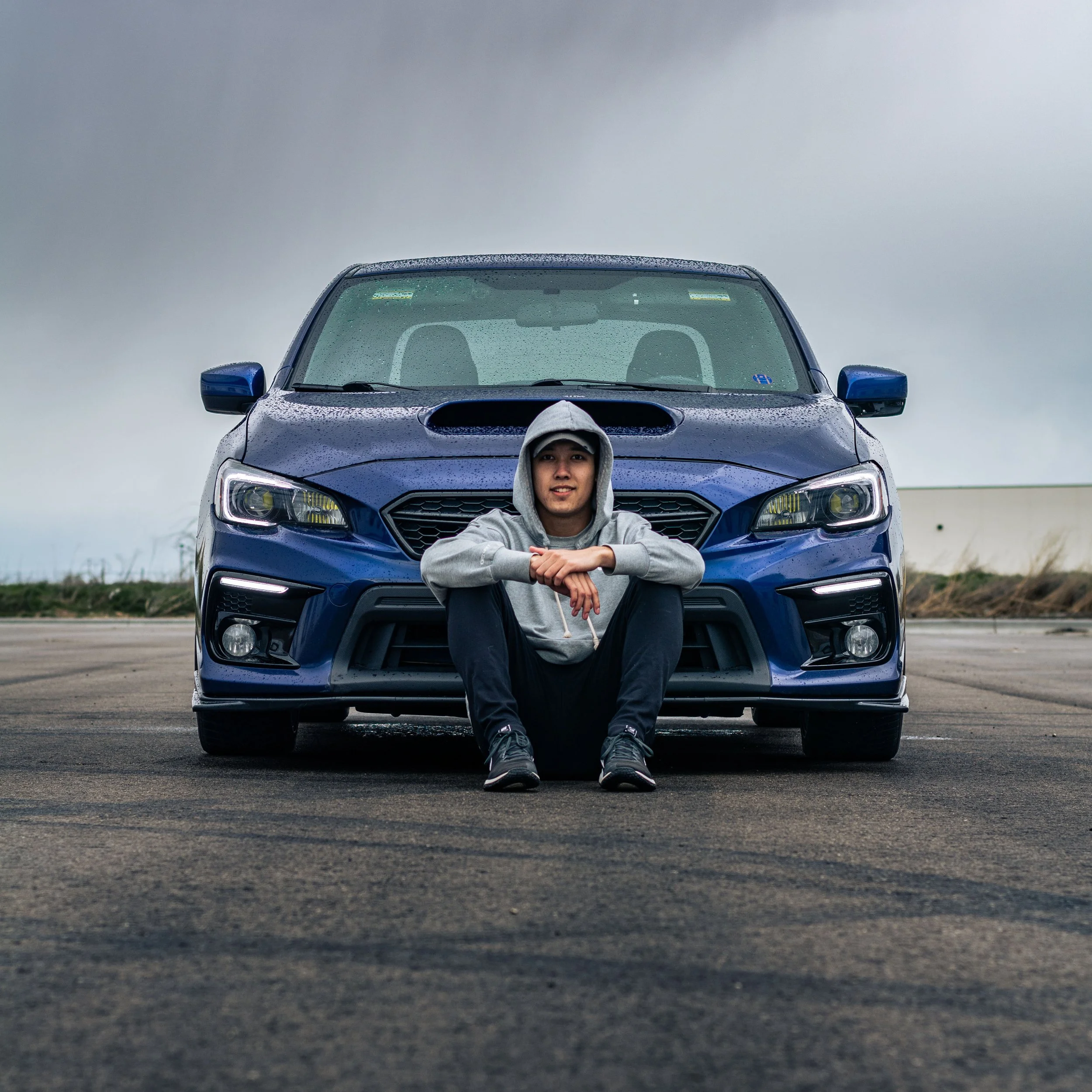 A person sitting on the ground in front of a blue car with water droplets on its surface, under a cloudy sky.