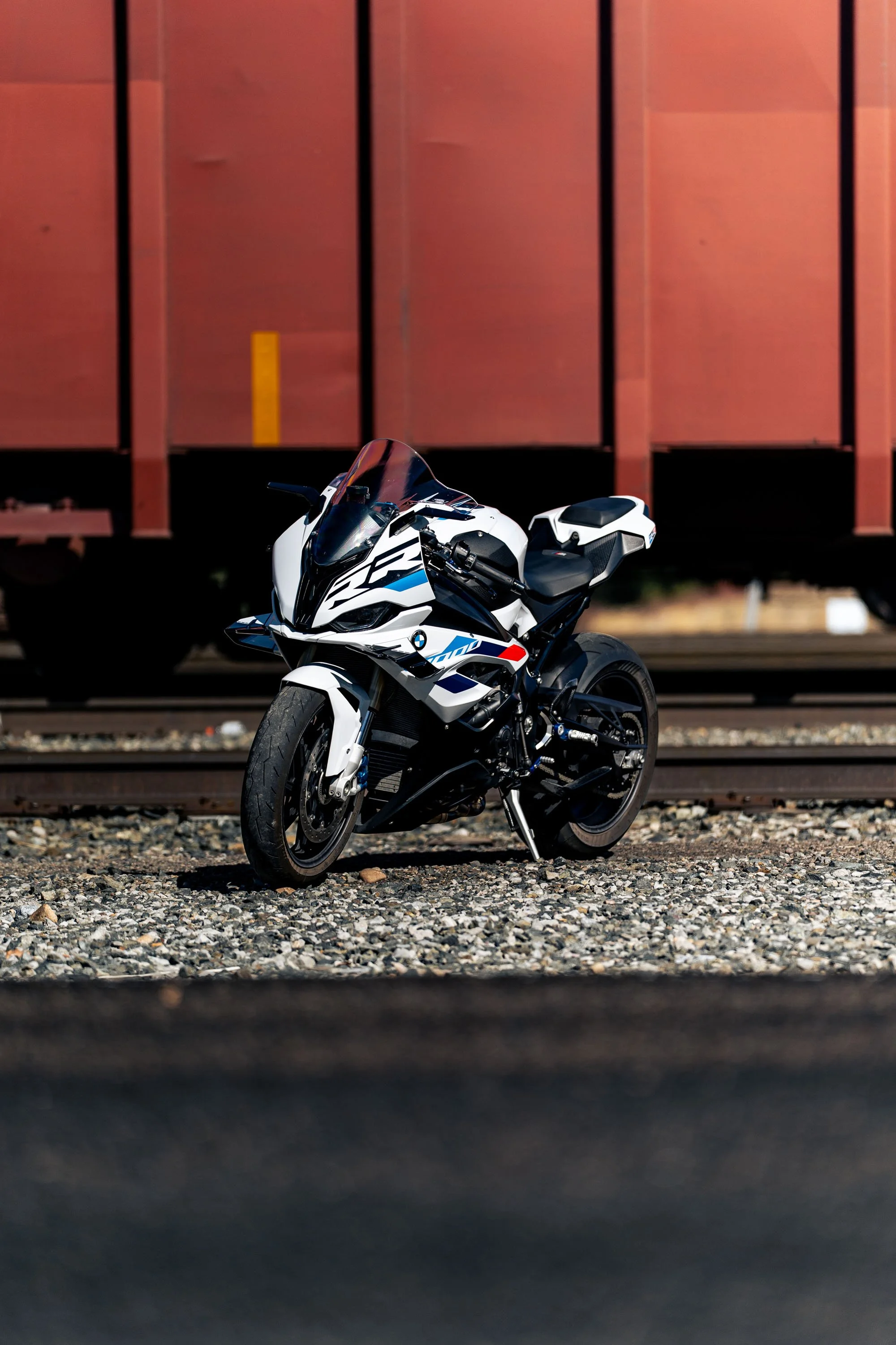 A white and black BMW sport motorcycle parked on a gravel surface in front of a red train car at a train yard.