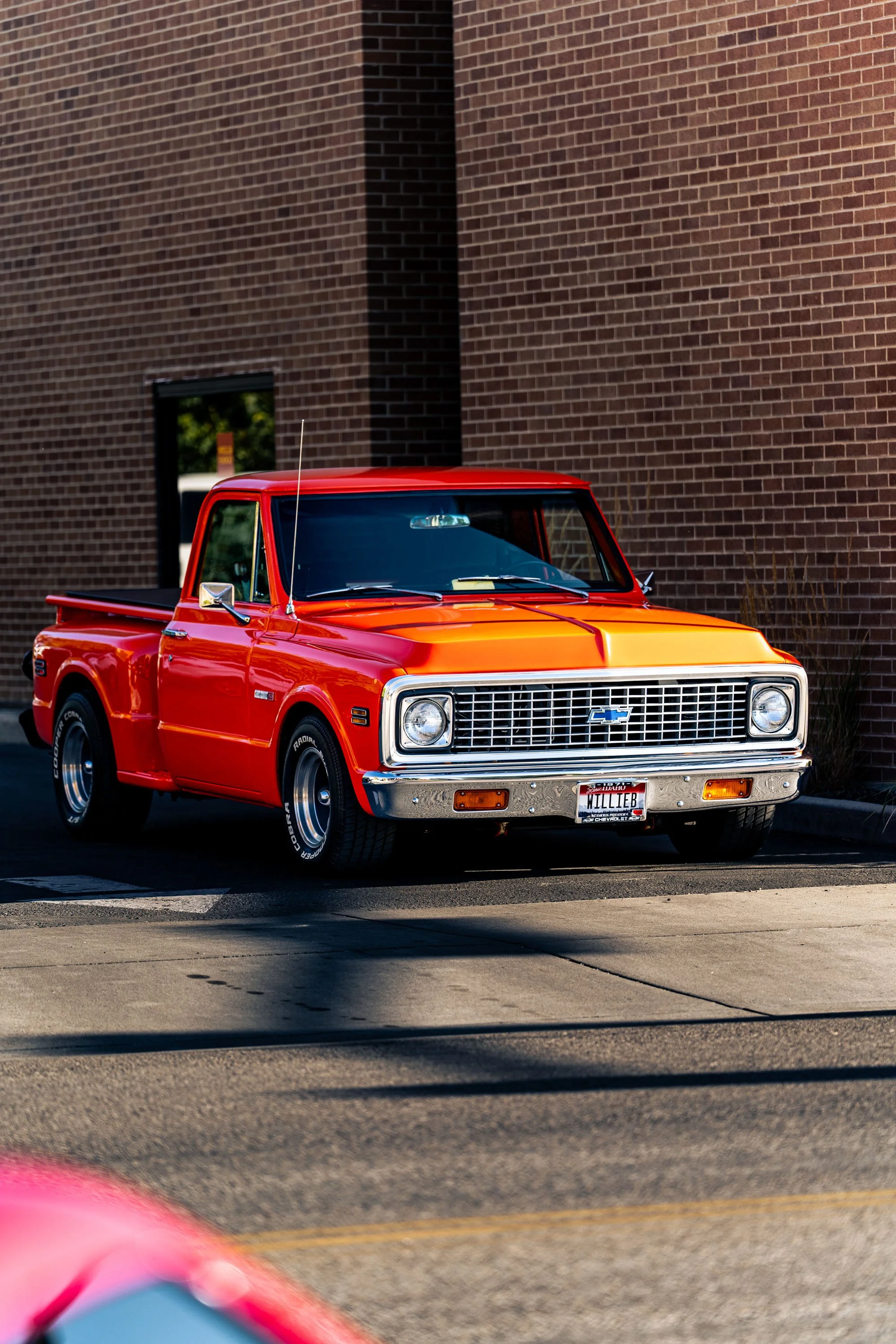 Red vintage pickup truck with a Chevrolet emblem on the grille, parked next to a brick building, with a blurred pink vehicle in the foreground.