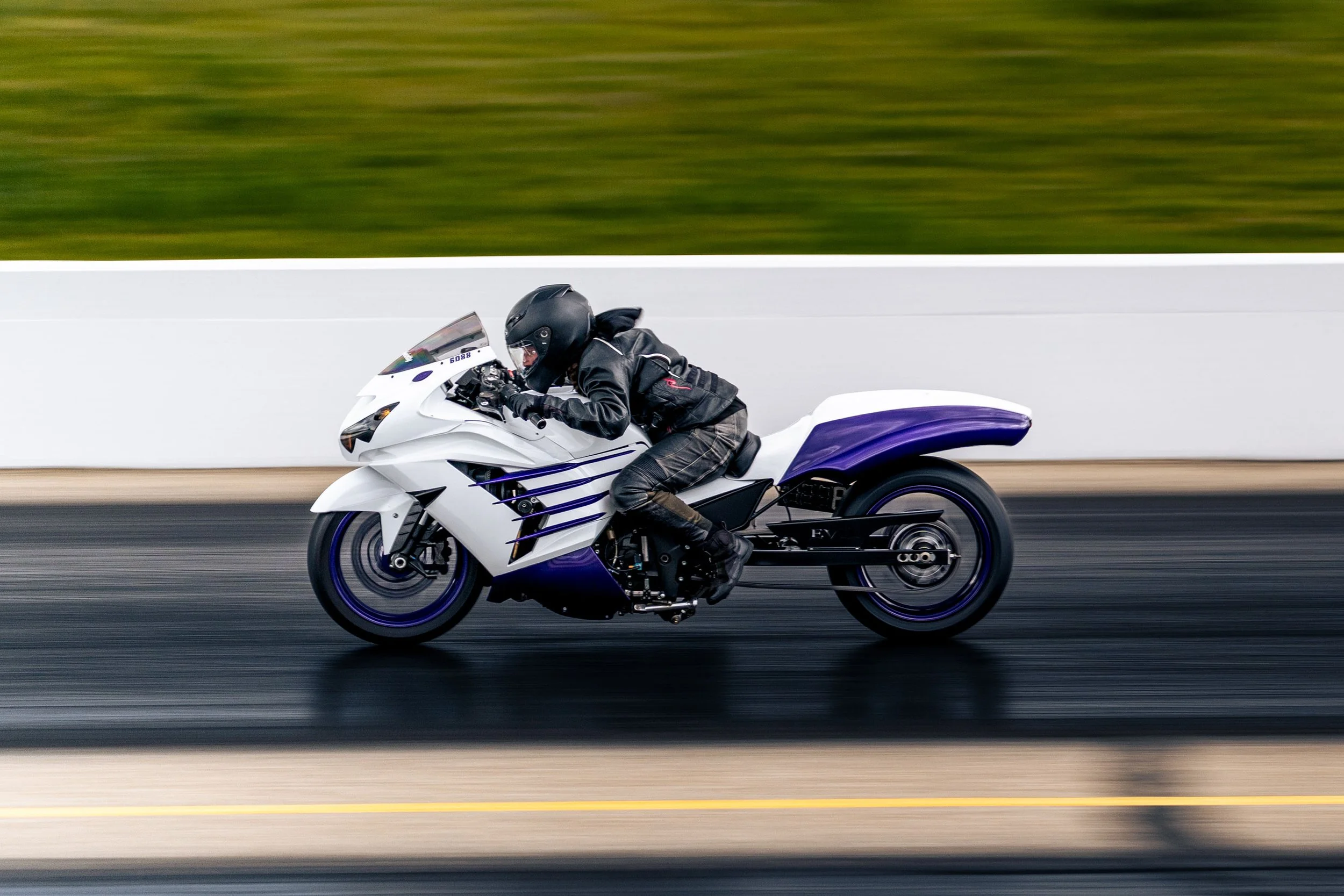 A person wearing a black helmet and leather jacket riding a white and purple motorcycle at high speed on a racetrack.