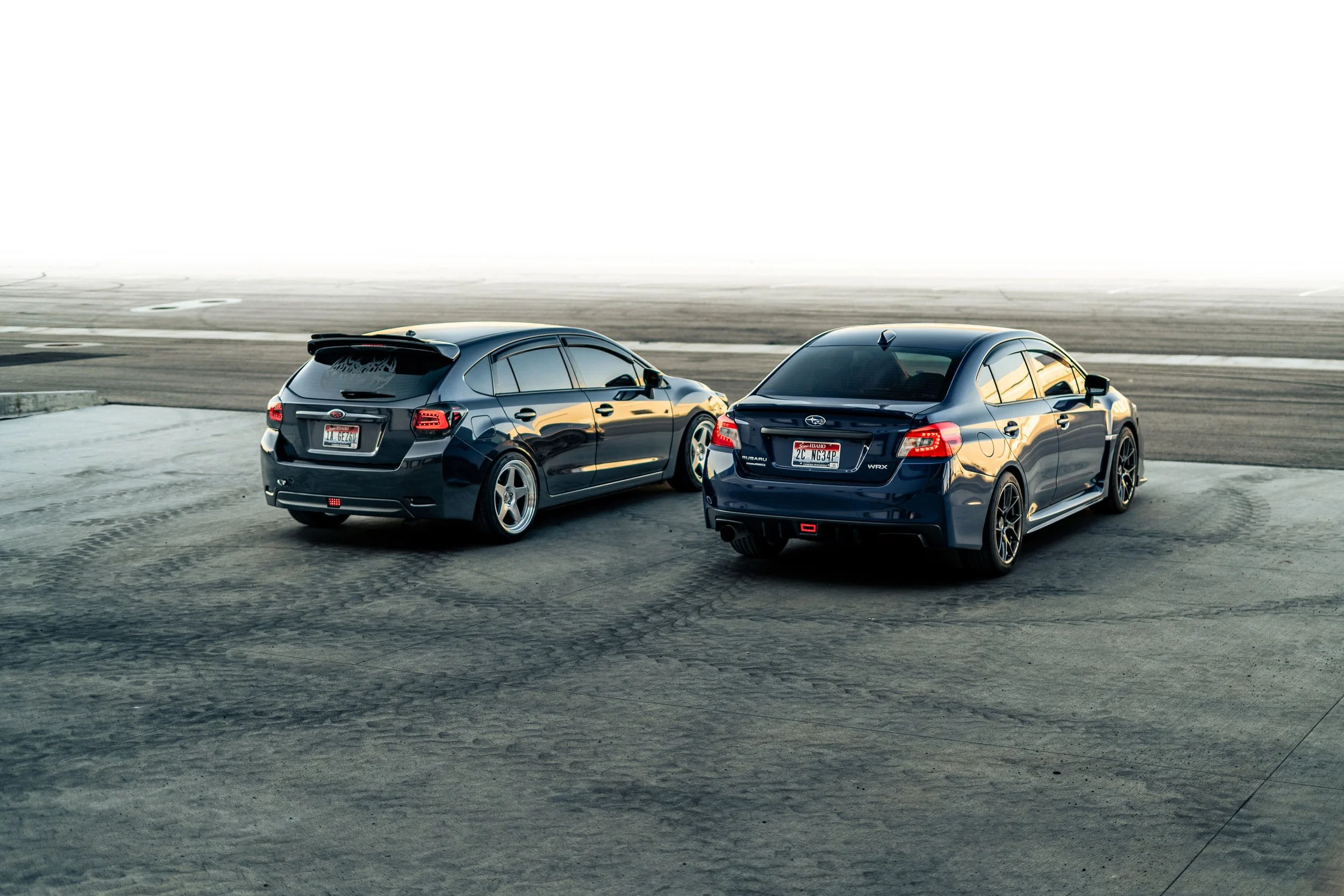 Two dark-colored Subaru cars parked on a concrete surface at a racing track with tire marks, under a clear sky.
