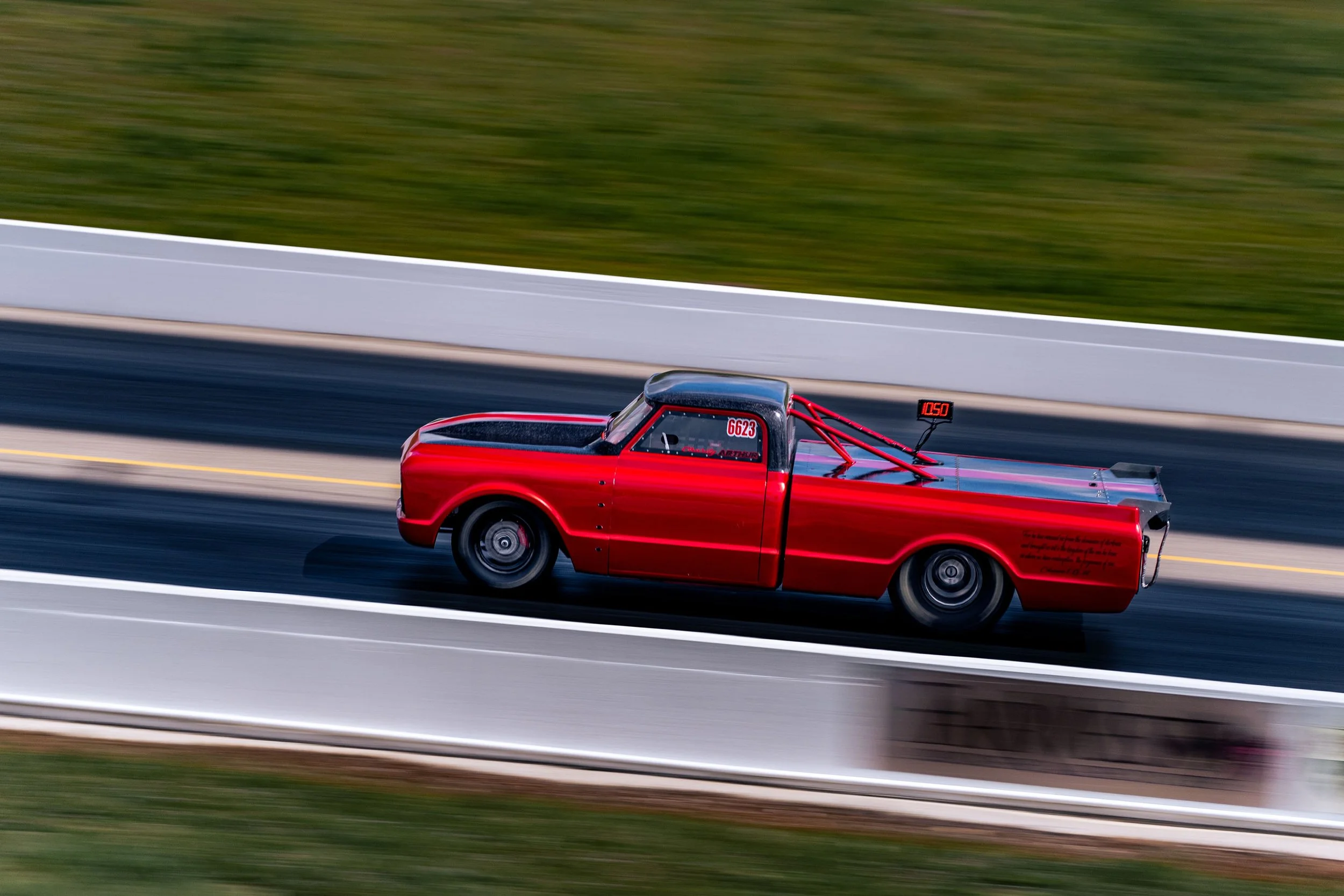 Red drag racing pickup truck with custom safety roll cage, moving at high speed on a racetrack with blurred background.