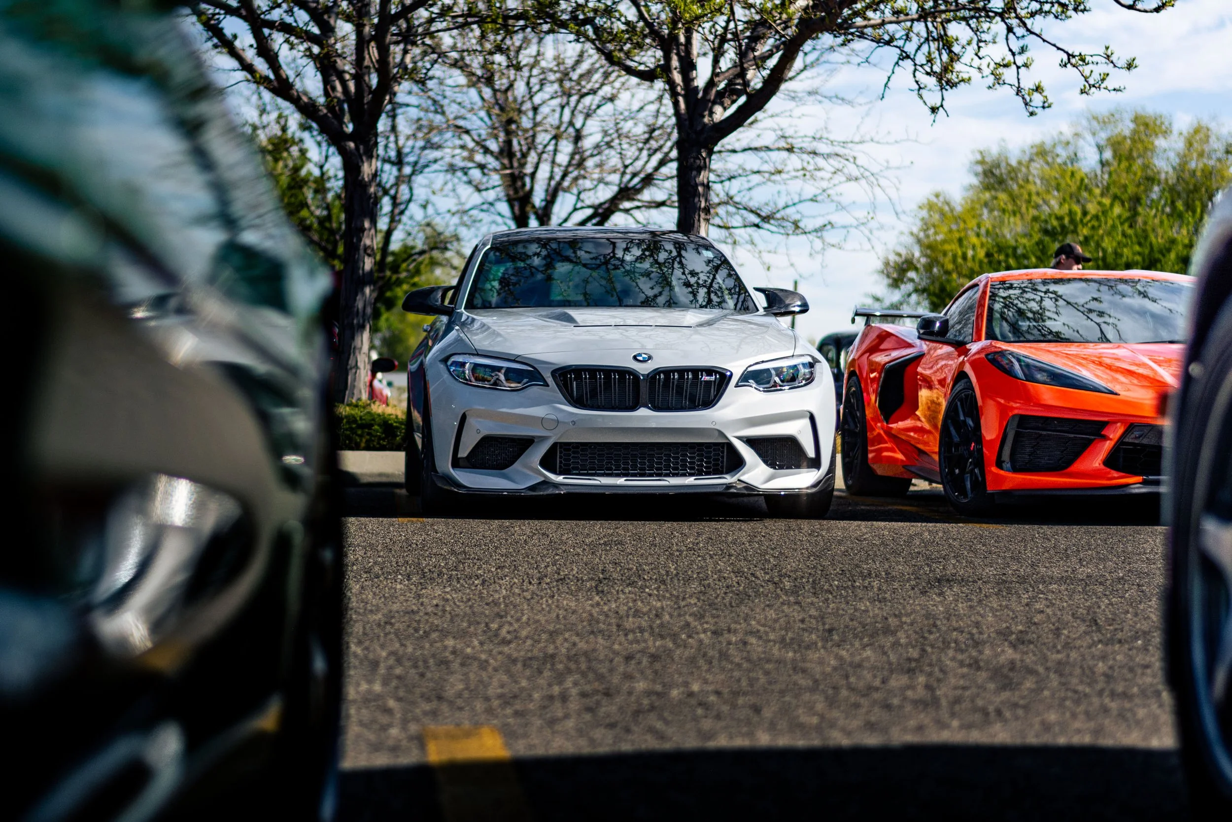 View of a parking lot with a white BMW and a red sports car parked among other vehicles, with trees and a cloudy sky in the background, seen from a low angle between two cars.