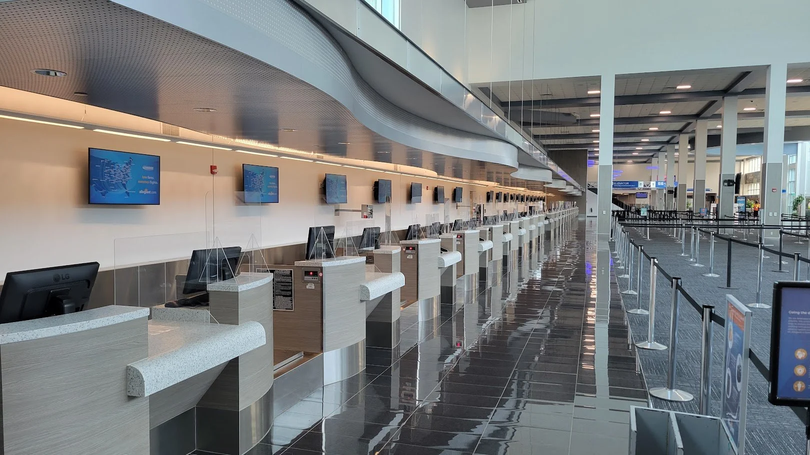 Empty check-in counters at an airport with television screens on the wall, stainless steel accents, and a reflective black tile floor.