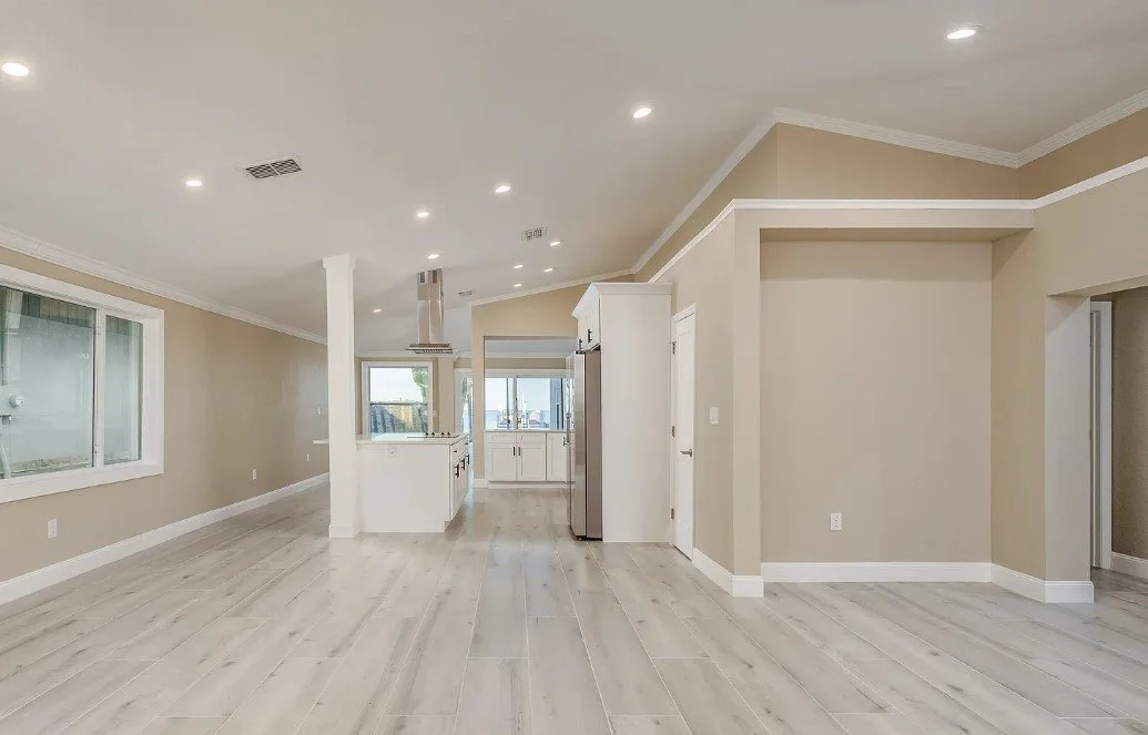 Empty open-concept living space with white flooring, beige walls, and white trim. Kitchen in background with white cabinets, stainless steel refrigerator, and windows overlooking outside. Recessed ceiling lights.
