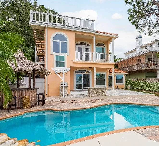 Backyard of a peach-colored, two-story house with a swimming pool in the foreground. The house features large arched windows, balconies with white railings, and a rooftop deck. There is a thatched-roof tiki bar on the left side.