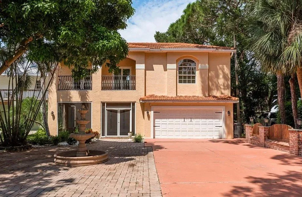 Two-story beige house with a red-tiled roof, two balconies, a large window, and a white garage door. The house features a brick driveway, surrounding greenery, and a fountain in the front yard.