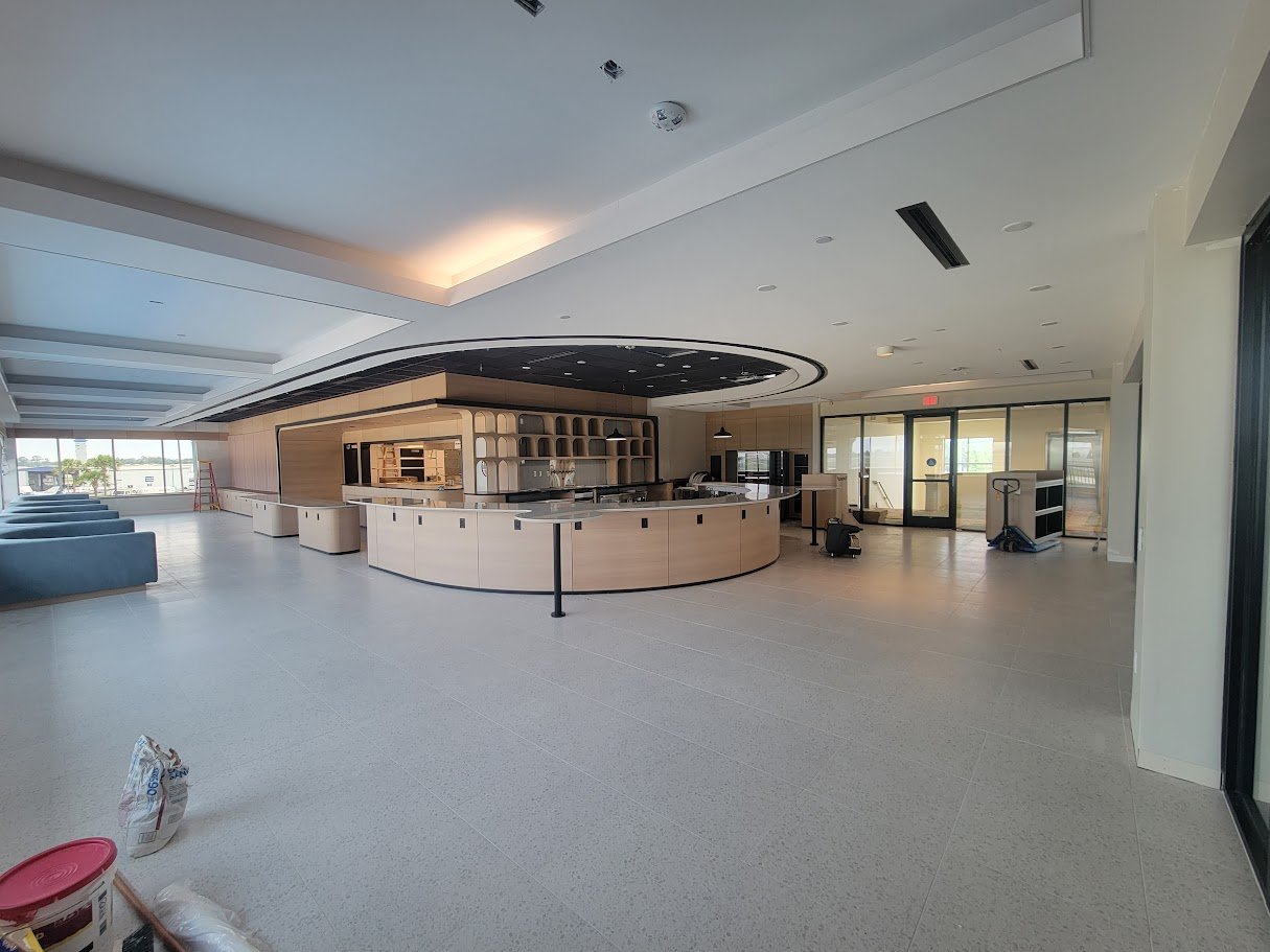 An empty modern lounge or lobby area with a curved wooden bar and open shelving, light-colored flooring, and large windows at the far end.