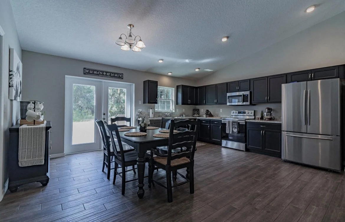 Kitchen with black cabinets, stainless steel appliances, a wooden dining table with six chairs, and a sliding glass door leading outside.