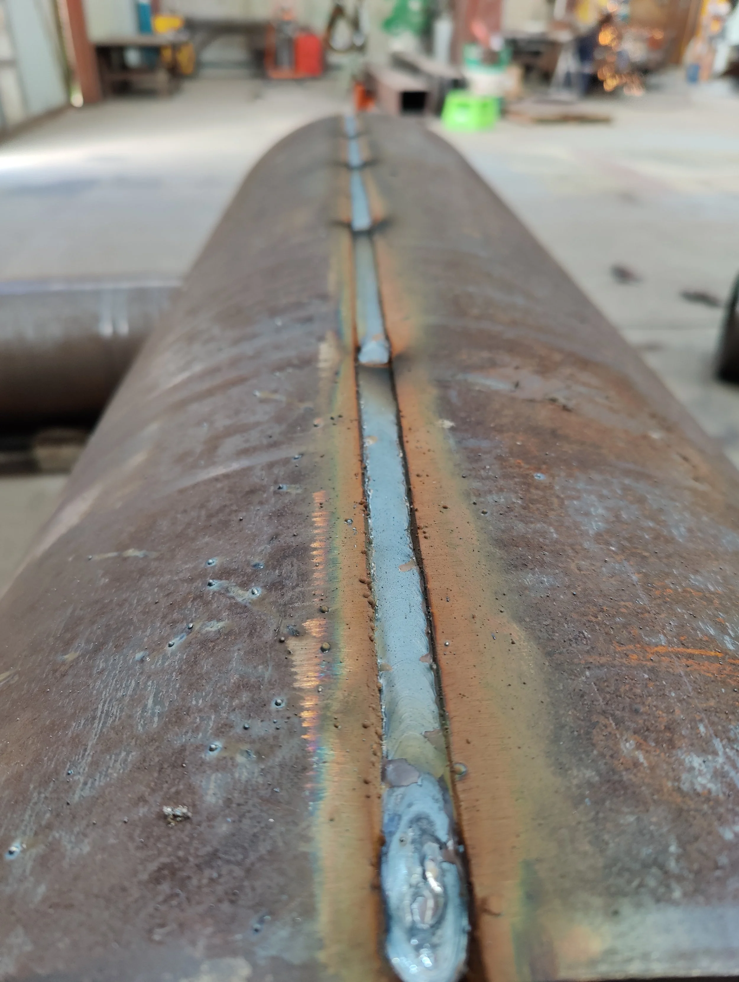 Close-up of a welded metal pipe in a workshop with blurred background of workbenches and tools.