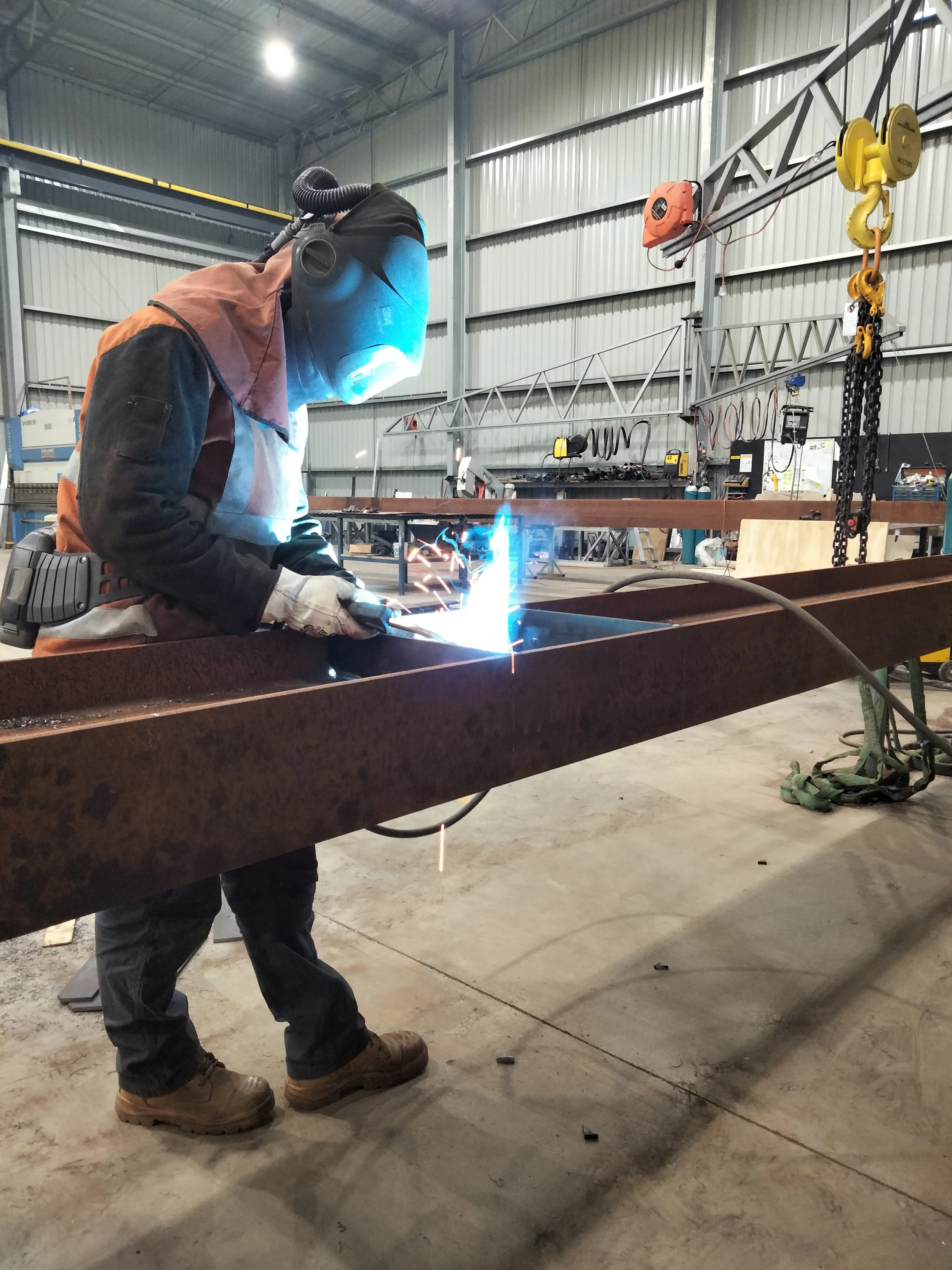 A worker wearing a welding helmet and protective gear is welding a metal beam in an industrial warehouse. Sparks and blue light from the welding process are visible, and various tools and equipment are mounted on the walls and ceiling.