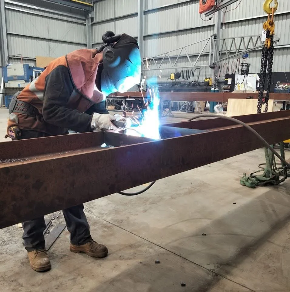 A person welding a metal beam inside a workshop, wearing safety gear including a welding helmet, gloves, and protective clothing.