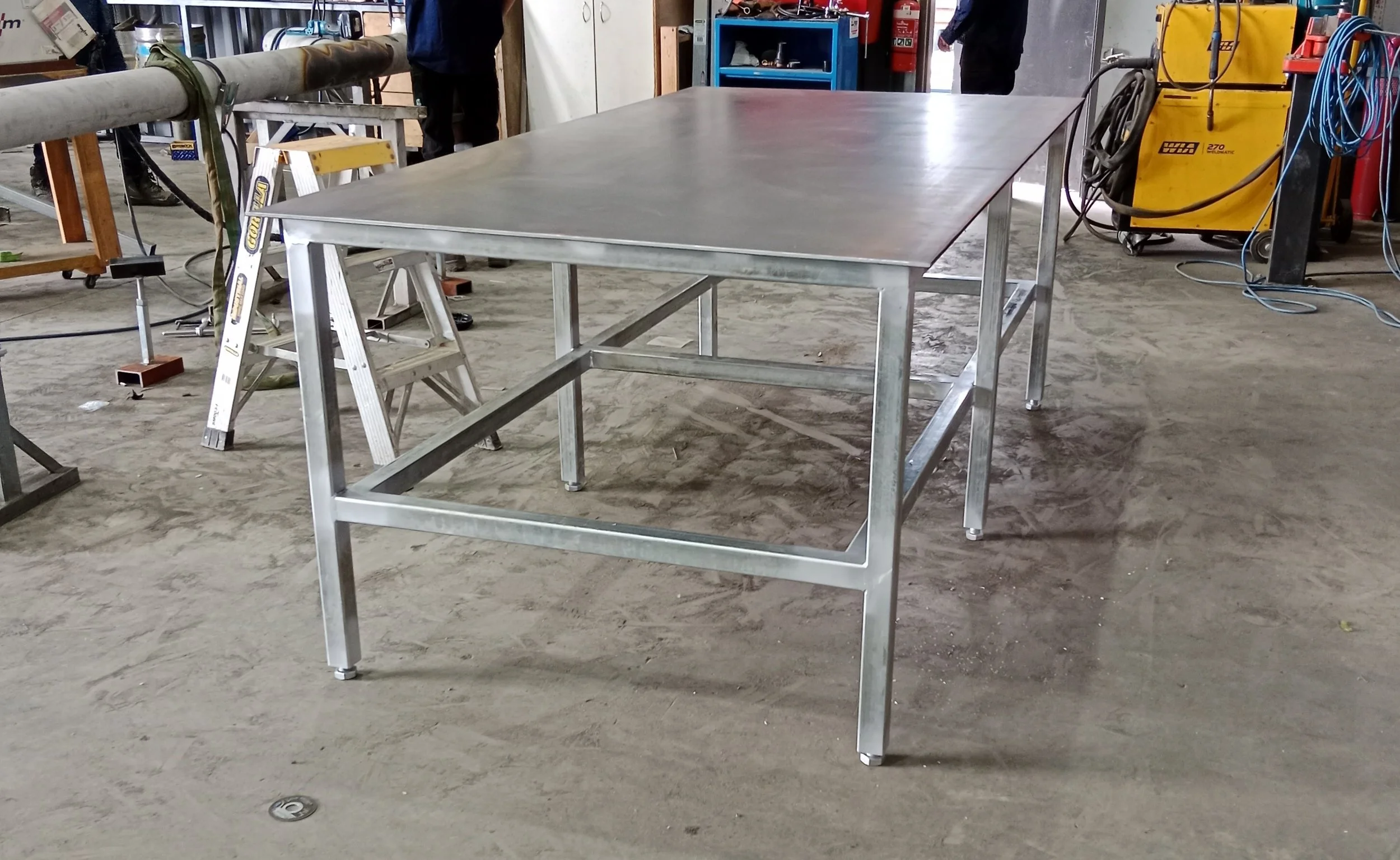 A metallic work table in a workshop or industrial setting, with some stairs and tools visible in the background.