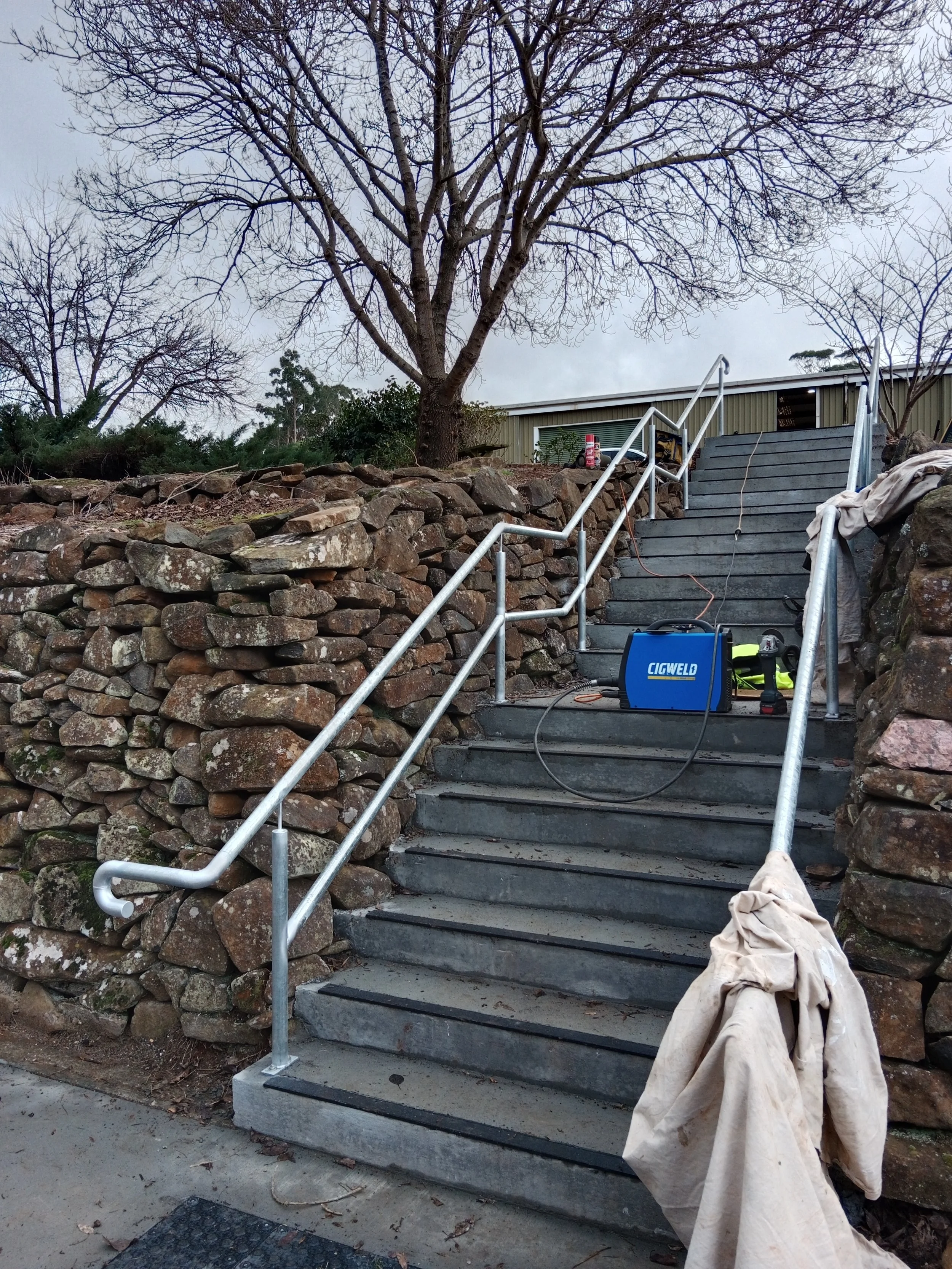 Construction site with outdoor staircase having metal railings, surrounded by stone walls and bare trees, with tools and equipment placed on and near the stairs.