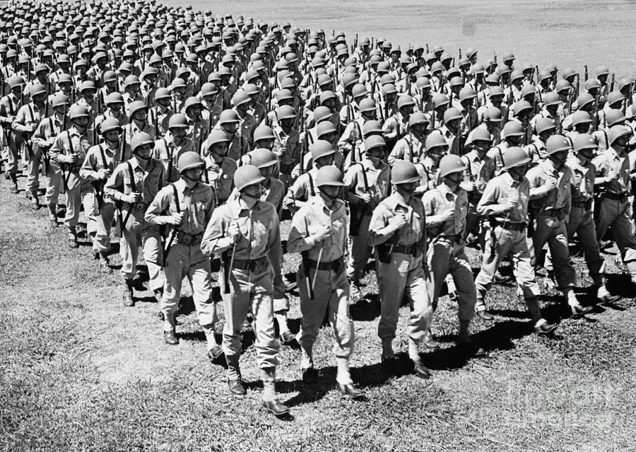 A large group of soldiers in uniform marching in formation on a grassy field, wearing helmets and carrying backpacks.