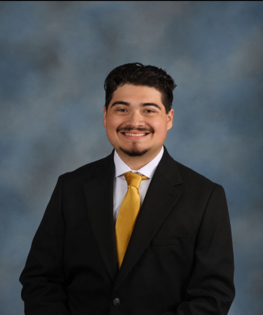 A young man with dark hair and a mustache, wearing a black suit, white shirt, and a yellow tie, smiling in a professional portrait against a blue-gray background.