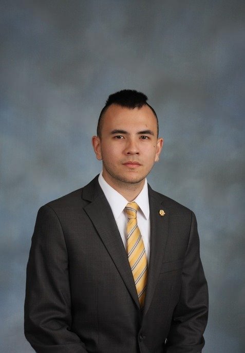 A young man with short black hair in a business suit and striped yellow tie, standing against a gray background.