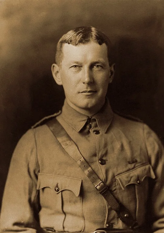 Black and white photograph of a man in military uniform with a strap across his chest, with short hair and a serious expression.