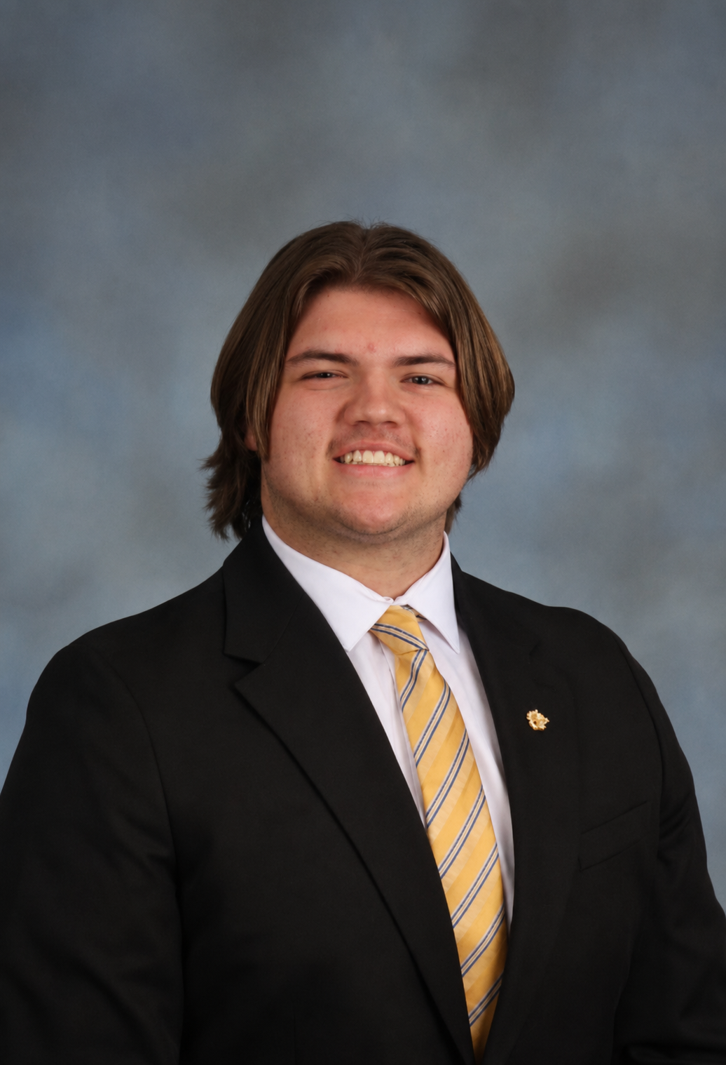 Young man in formal black suit, white shirt, and gold-striped tie, smiling against a gray background.