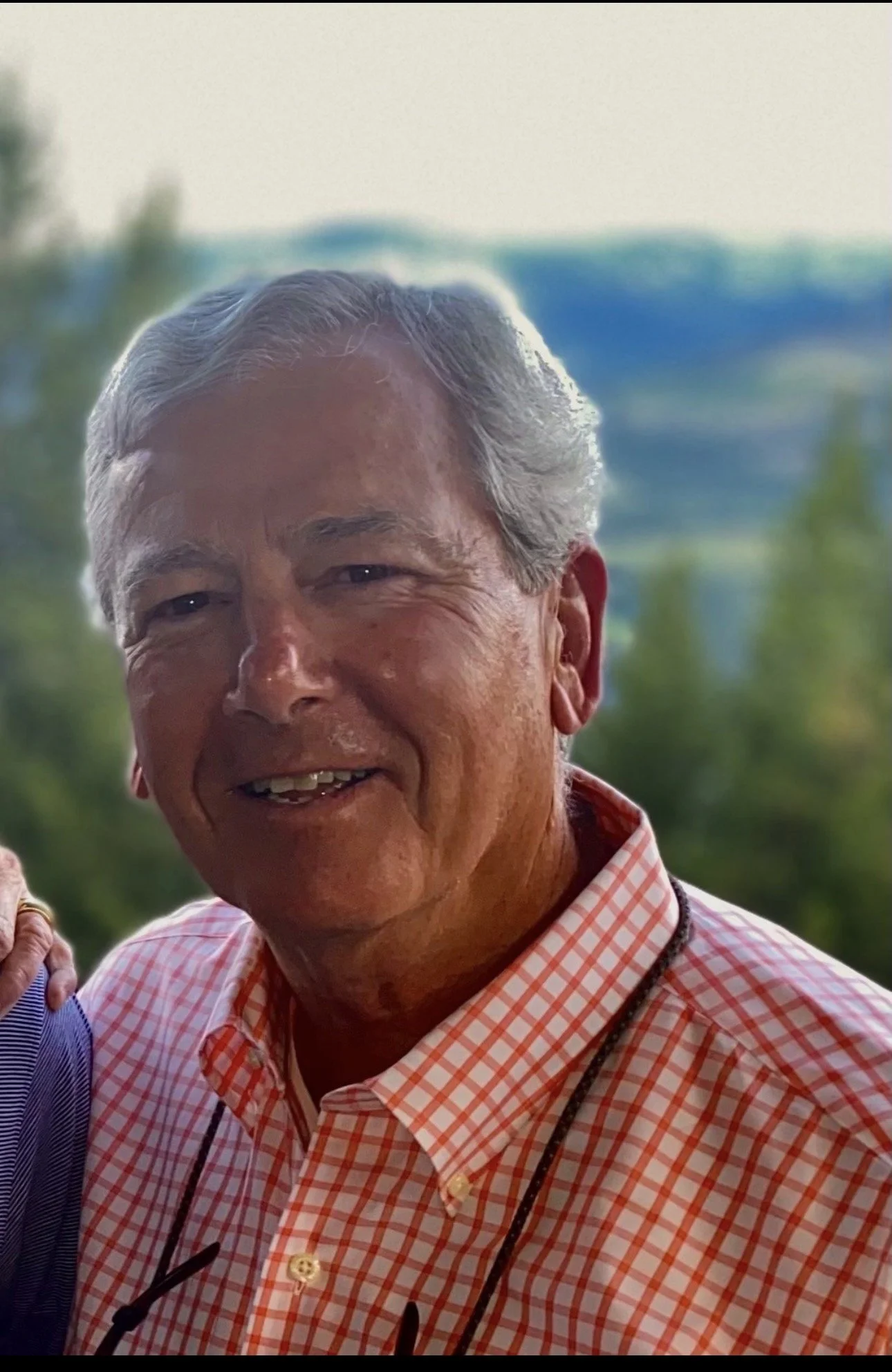 A smiling man with gray hair wearing a pink checkered shirt outdoors with blurred trees and a building in the background.