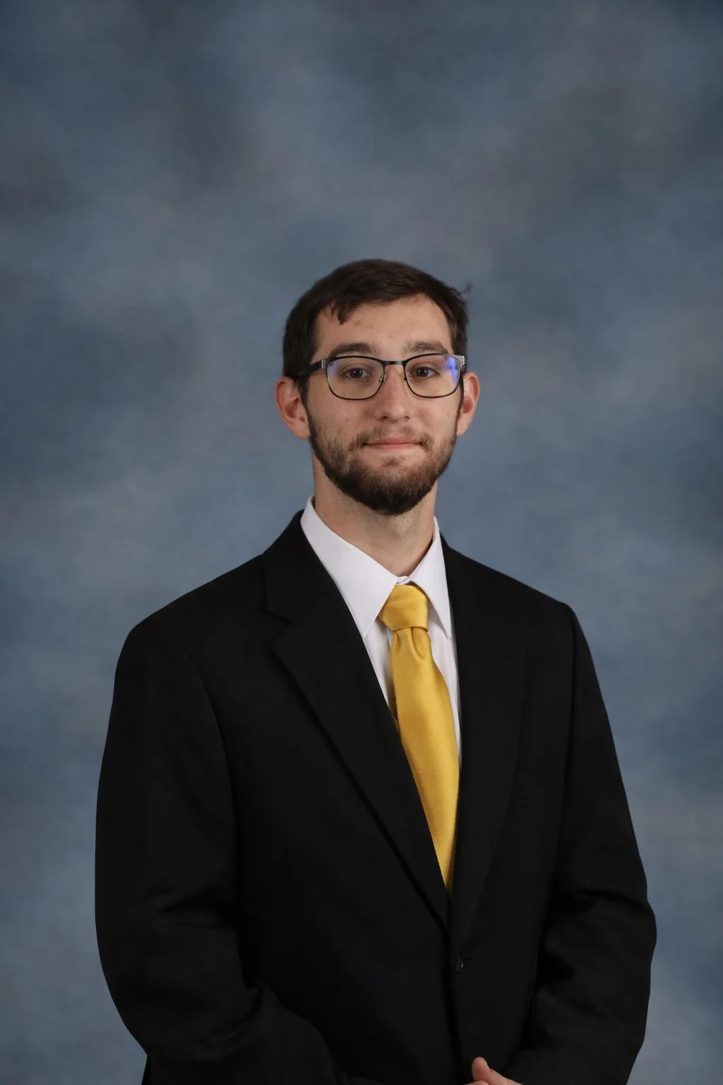 A young man wearing glasses, a black suit, white shirt, and yellow tie posing against a blue-gray gradient background.