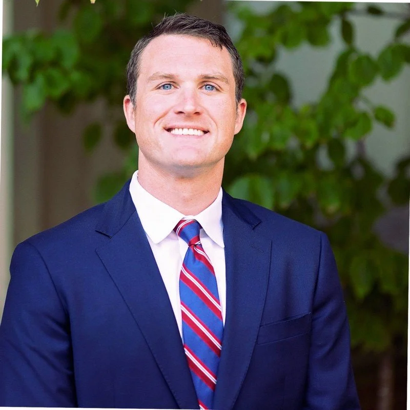 A man in a dark blue suit, white shirt, and a striped tie smiling outdoors with green foliage in the background.