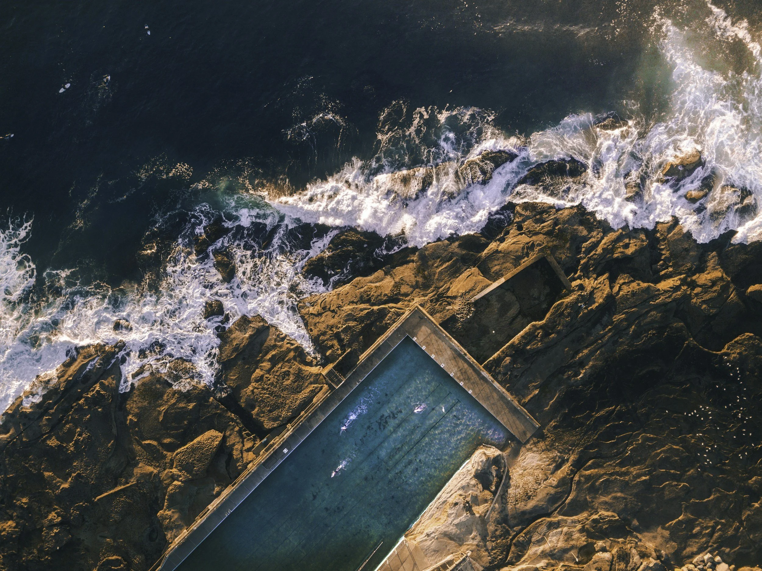 Overhead view of waves crashing against rocky shoreline with a pool in the foreground.