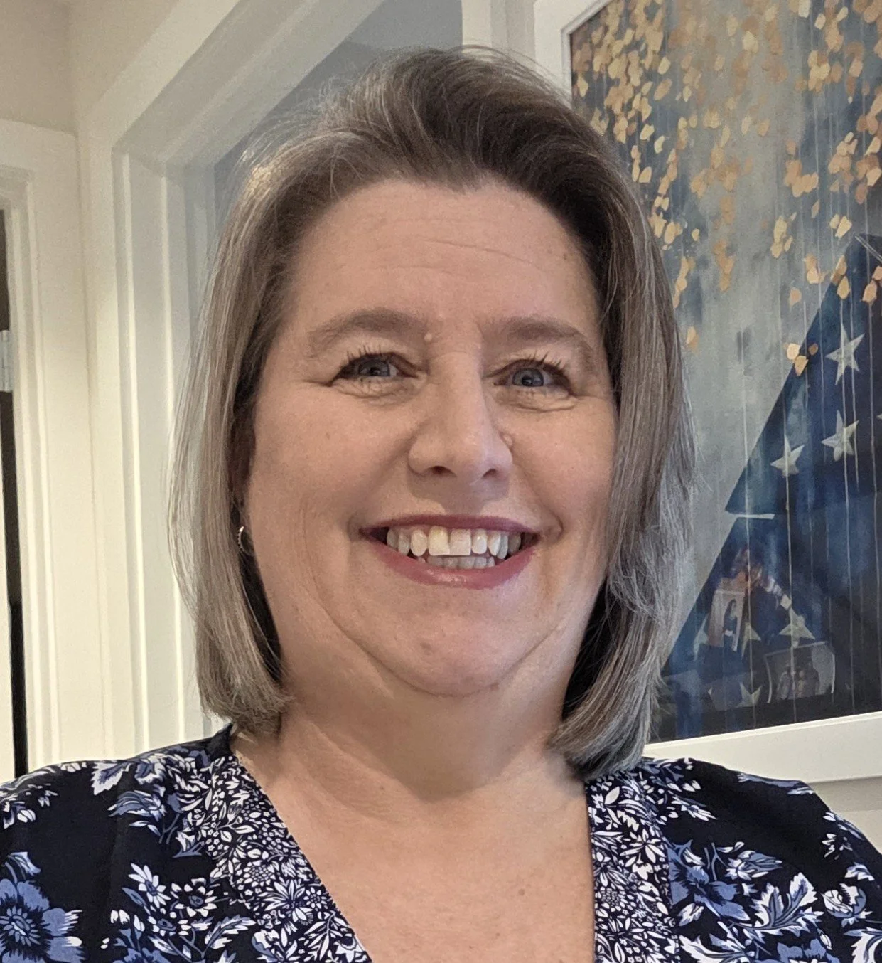 A woman with shoulder-length gray hair smiling shows her teeth, wearing a blue floral patterned top, standing indoors with a patriotic or star-themed decoration in the background.