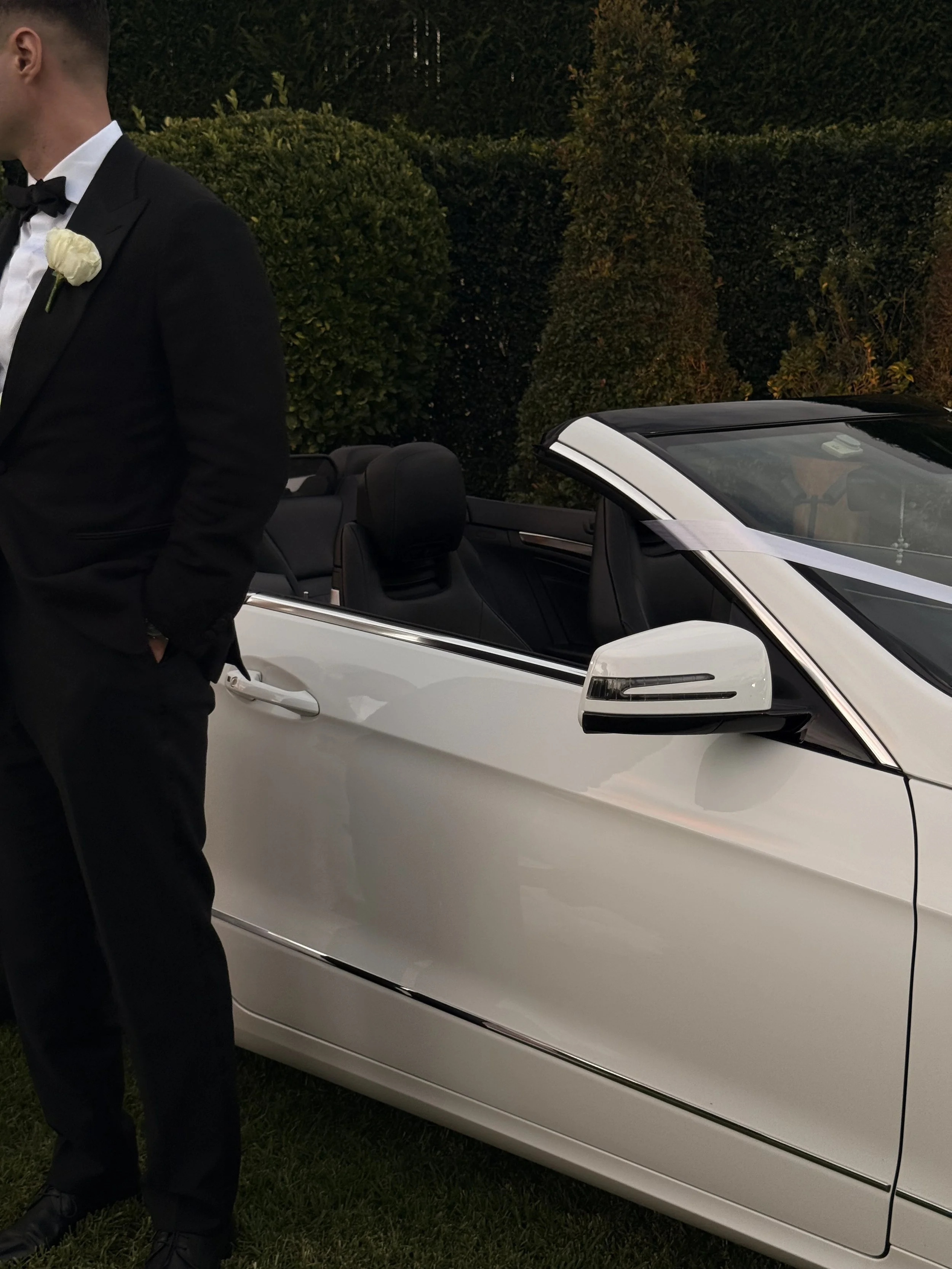 A groom in a tuxedo with a white boutonniere standing next to a white convertible car with black interior, located at Miramare Gardens, Sydney.