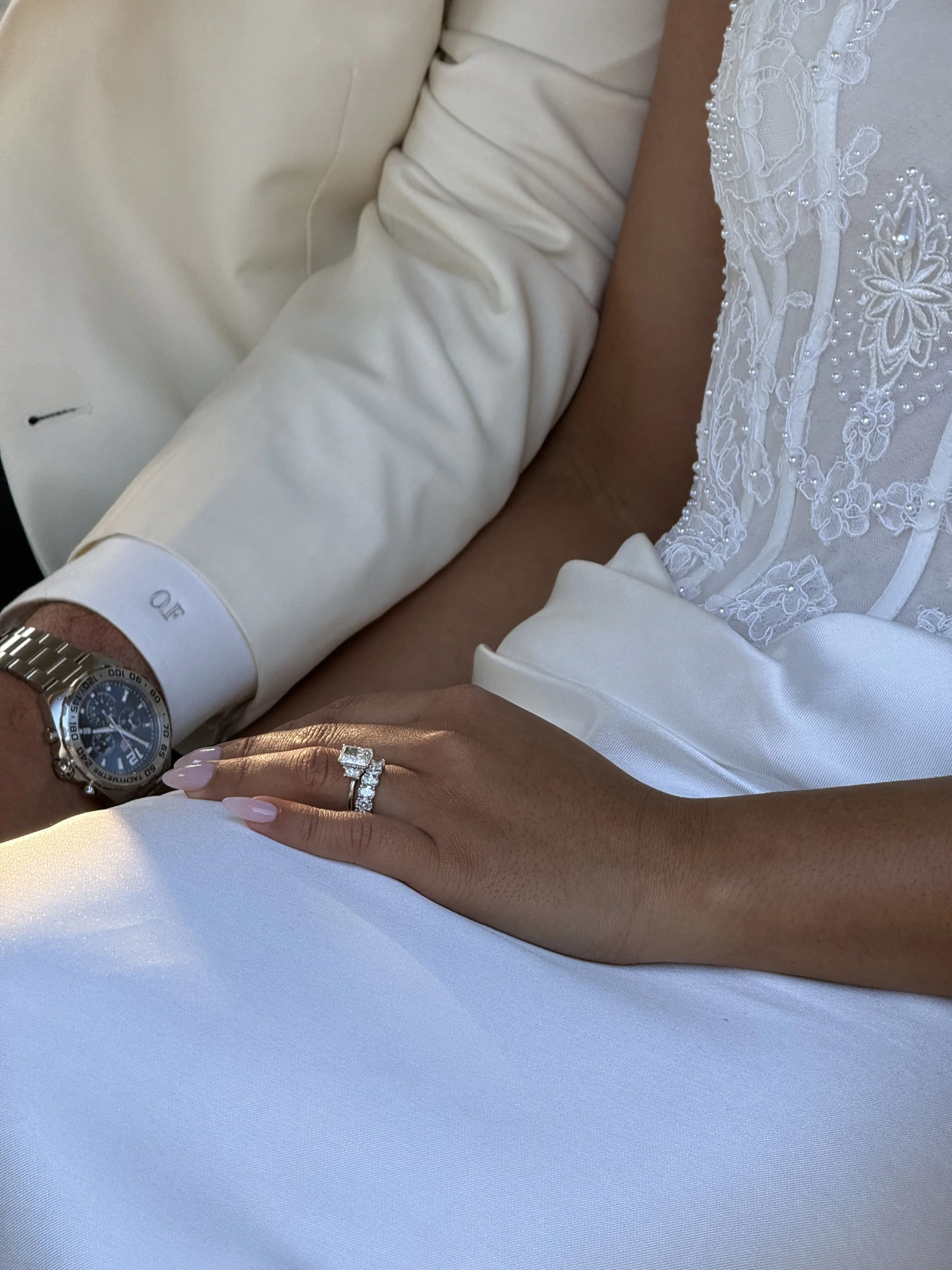 Close-up of a couple's hands with wedding rings against the woman's wedding dress. The woman is wearing a lace wedding dress with bead embroidery, and the man is wearing a white shirt with a wristwatch.