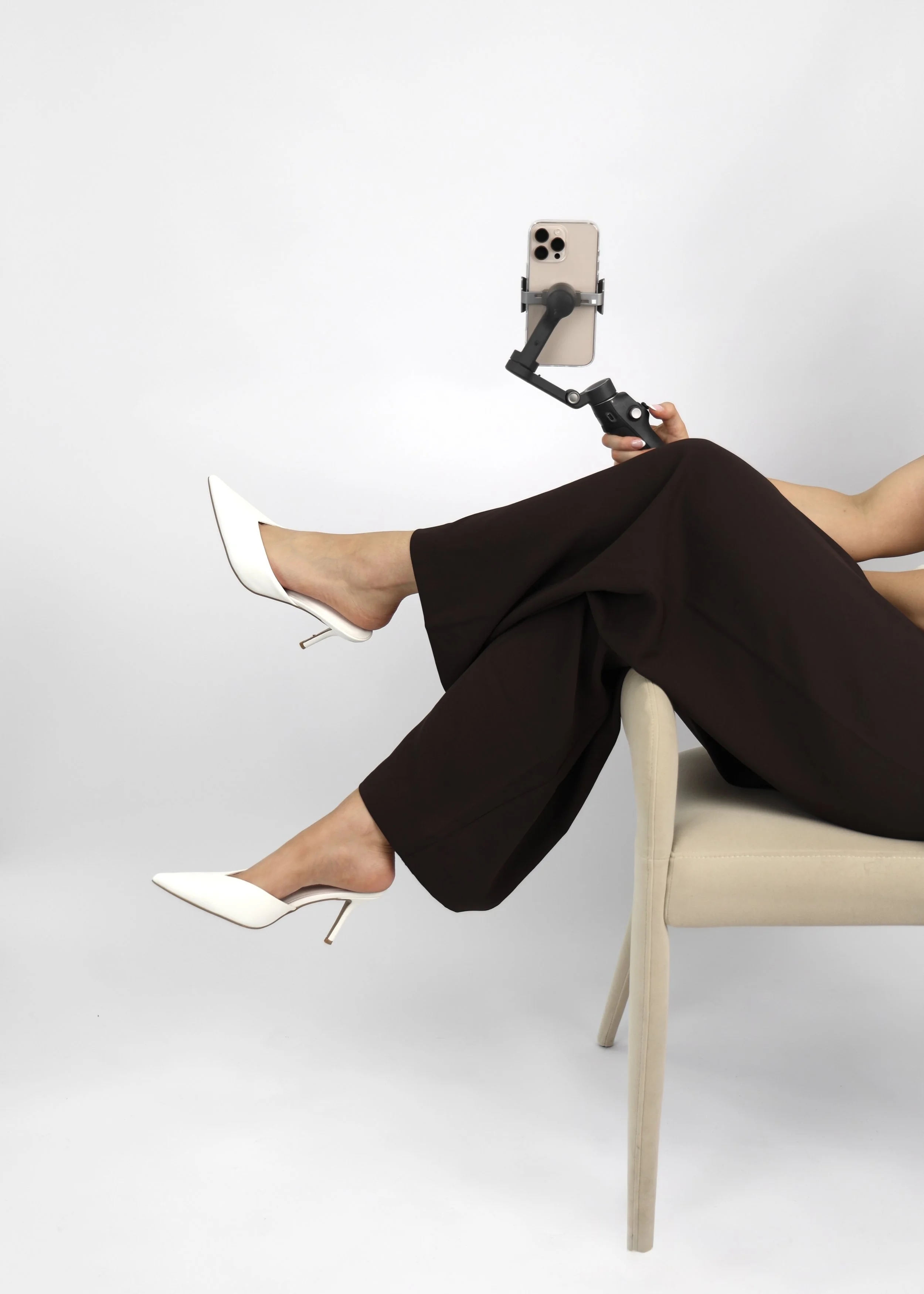 The founder with crossed legs wearing white high heels, chocolate brown pants, holding an Iphone on a stabilizer, sitting on a light-colored chair against a white background.