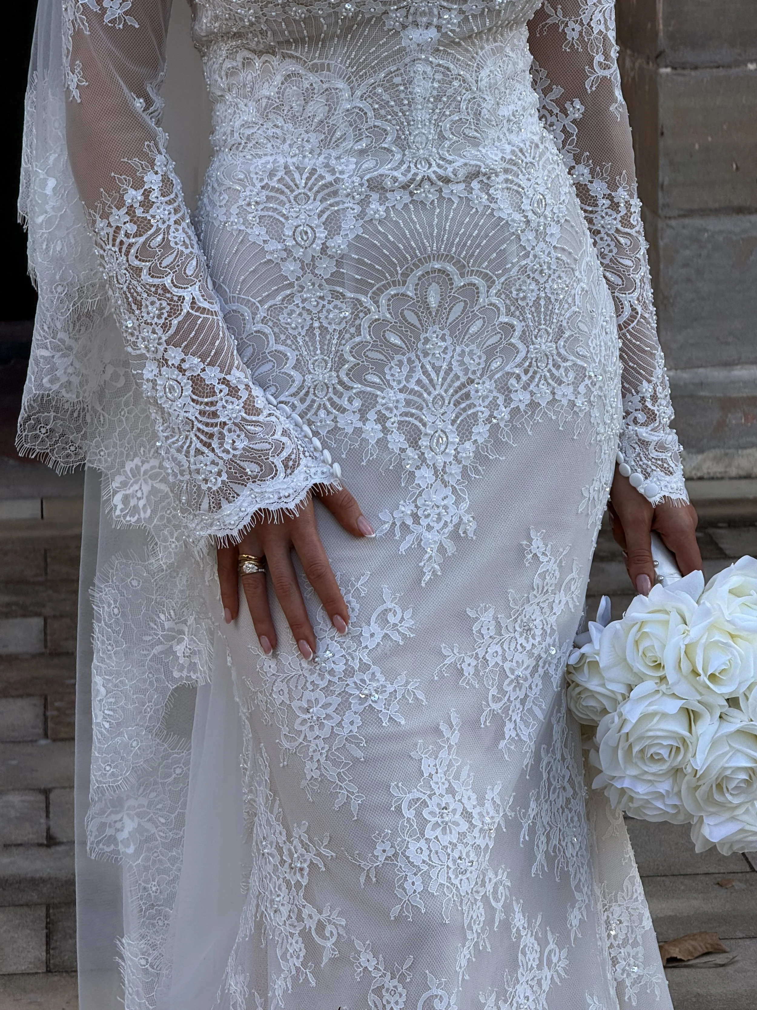 Close-up of a bride in a white lace wedding dress with floral and geometric patterns, holding a bouquet of white roses.