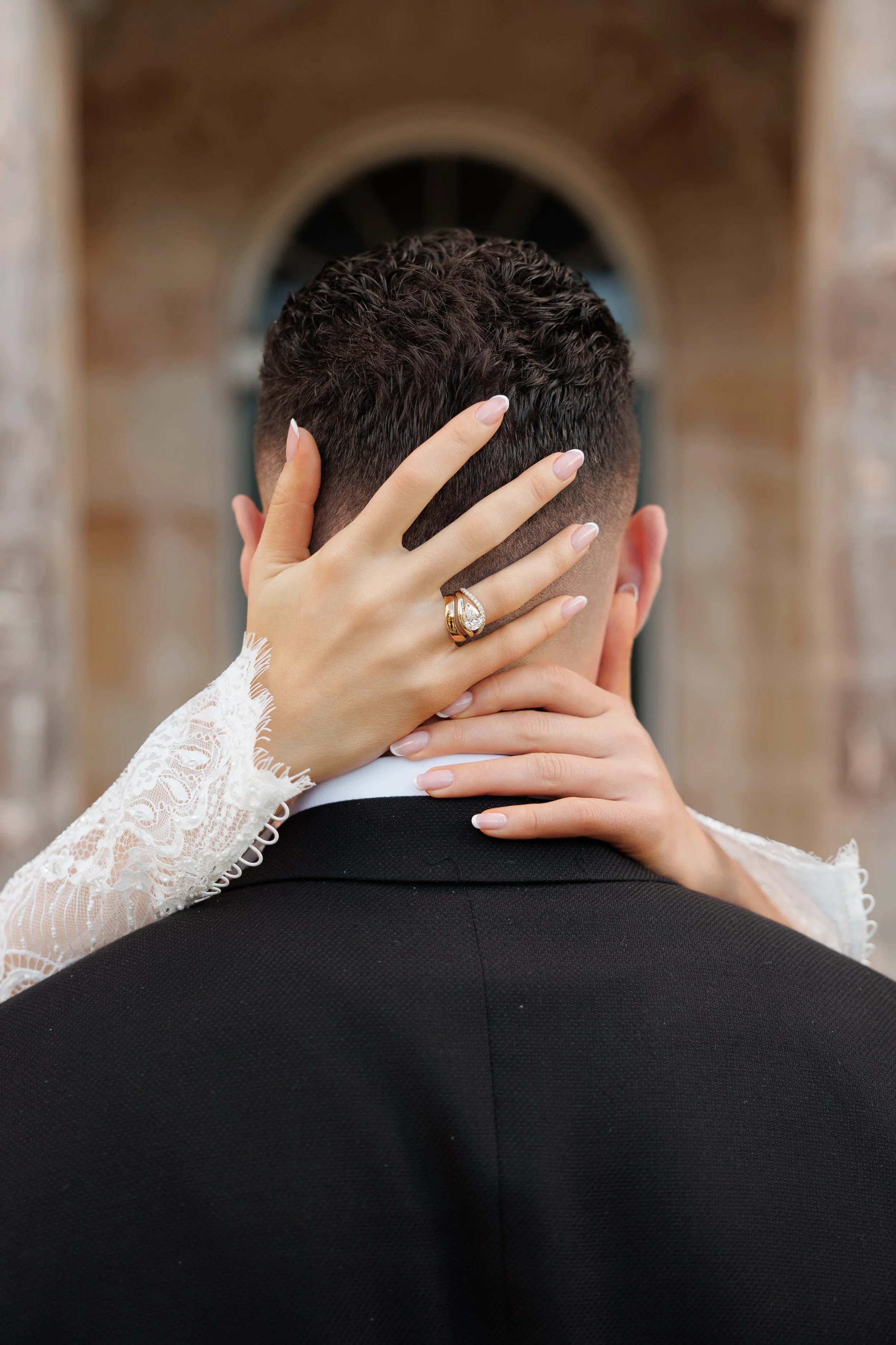 Close-up of a bride and groom, with the bride's hand on the back of the groom's head, showing her wedding ring. The bride is wearing a lace sleeve. The groom is dressed in a black suit, and the scene is outdoors near a brick building.