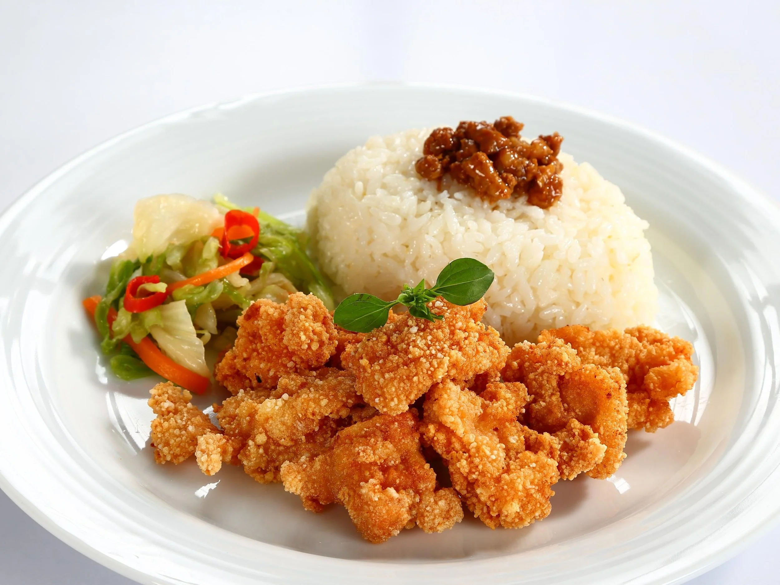 Fried chicken pieces, a scoop of white rice with a topping of meat sauce, a small serving of salad with lettuce, shredded carrots, and red chili slices, garnished with fresh basil leaves, all on a white plate.