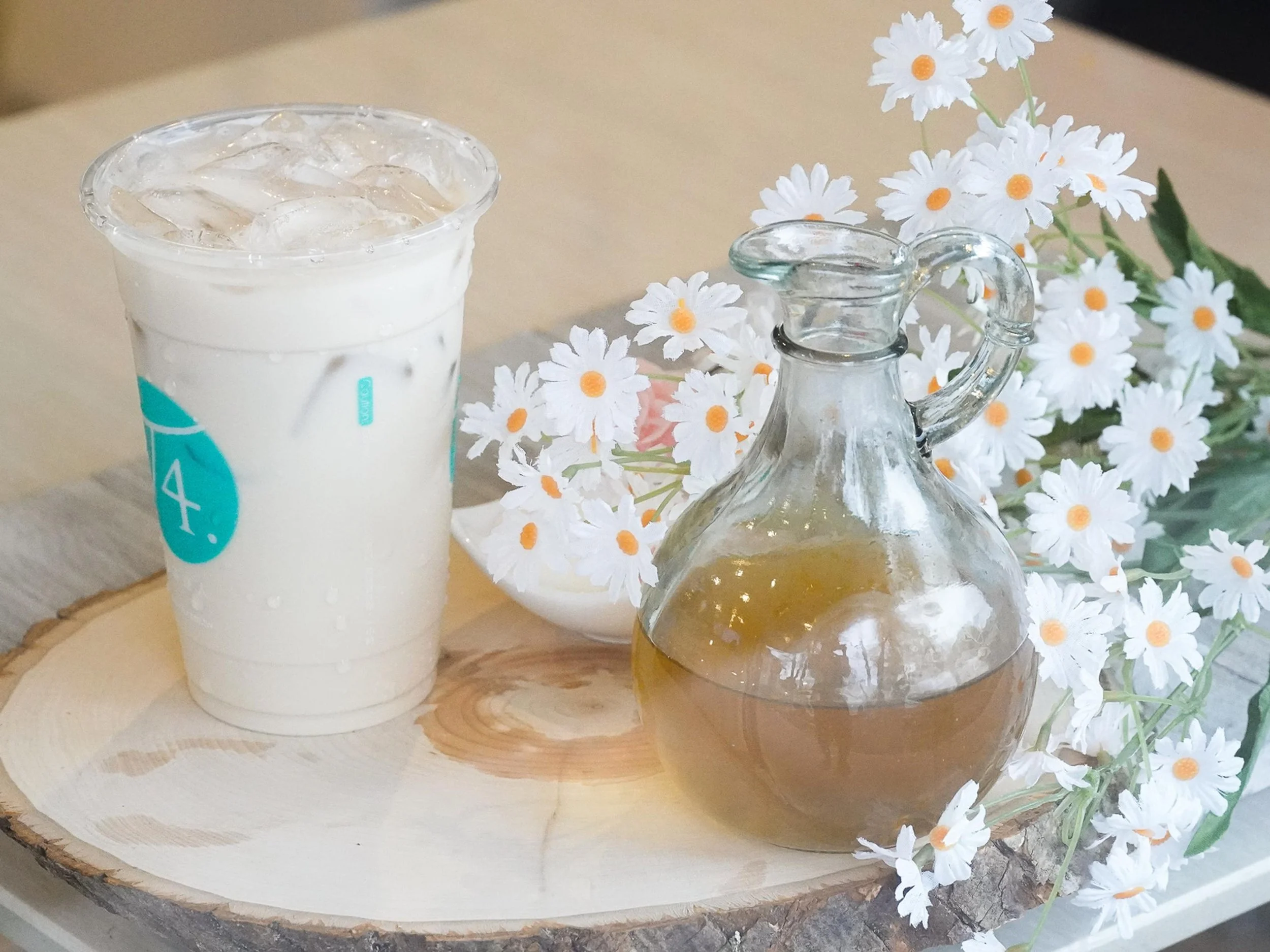 Glass pitcher and a cup filled with a light-colored beverage, barley, placed on a round wooden tray decorated with white daisies and greenery.