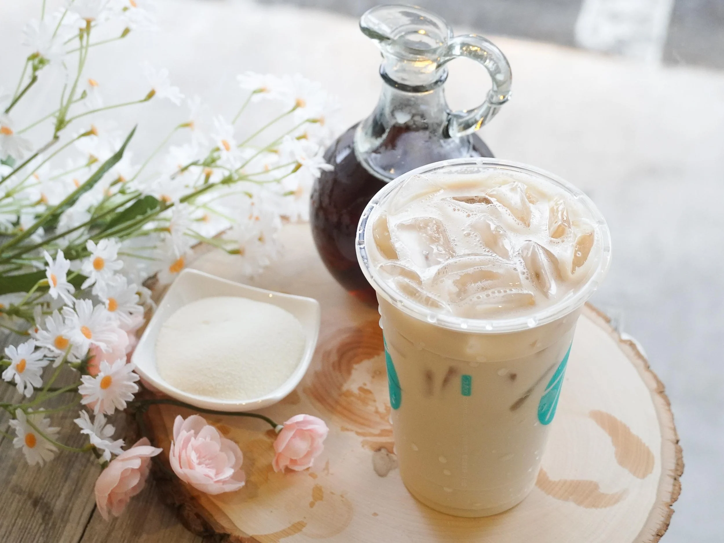 A transparent glass pitcher filled with dark liquid, possibly iced coffee, is in the background. In the foreground, there's a clear plastic cup with a cold, creamy iced beverage topped with ice. To the left, there are two small white bowls, one fille