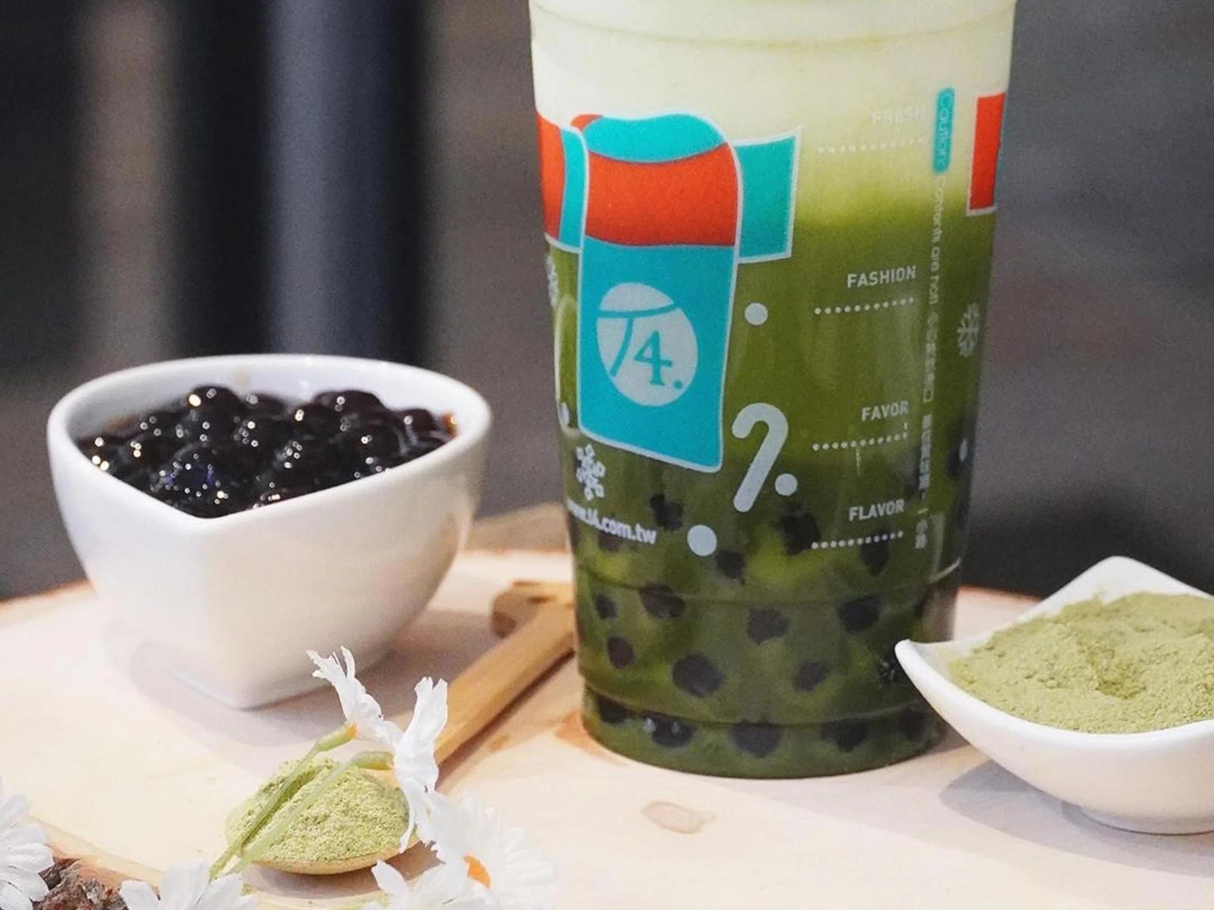 A cup of black bubble tea, a green bubble tea drink with tapioca pearls, and bowls of matcha and green tea powders on a table.