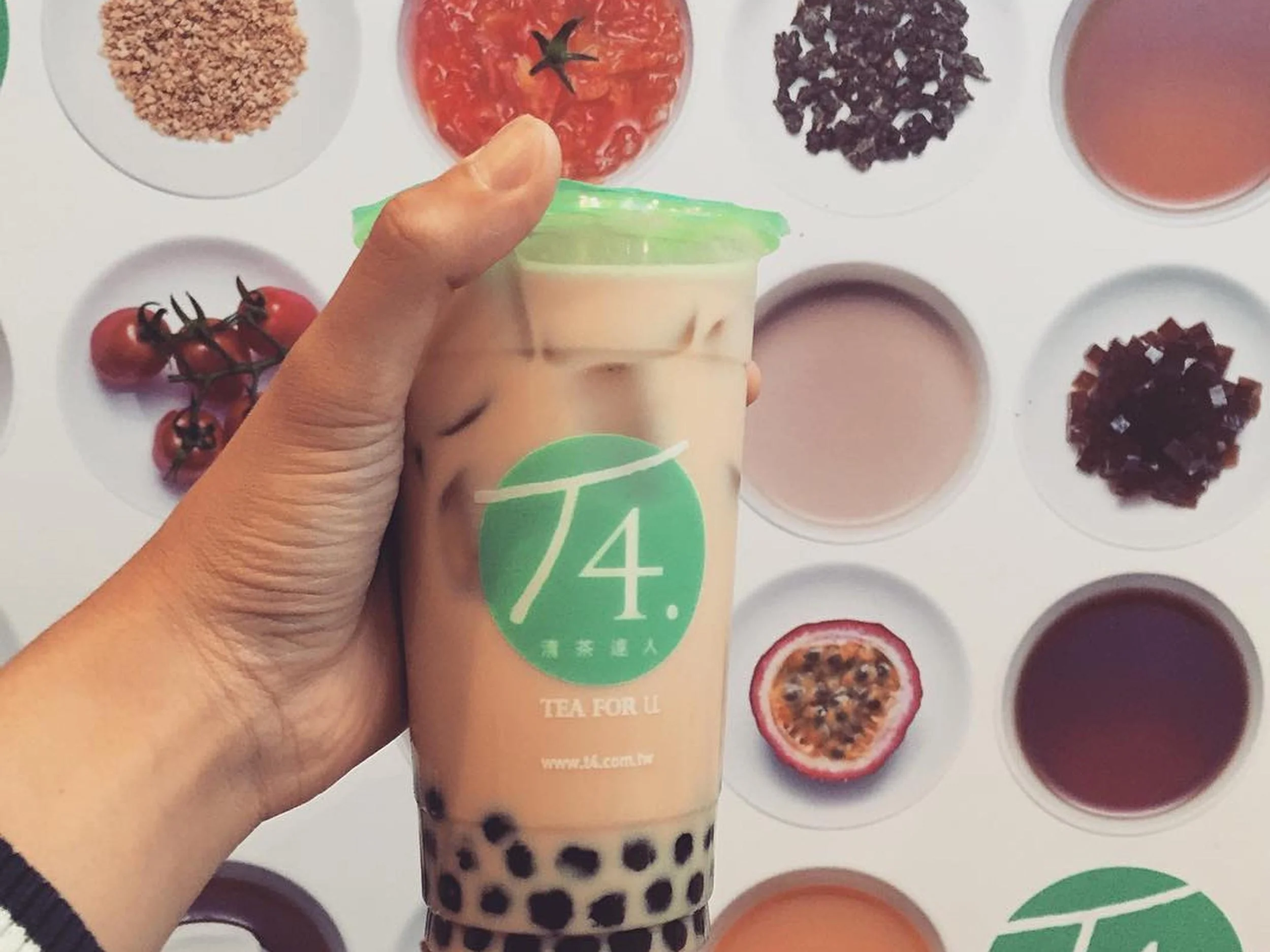 A person holding a container of bubble tea, with various toppings and ingredients in small dishes on a white surface in the background.